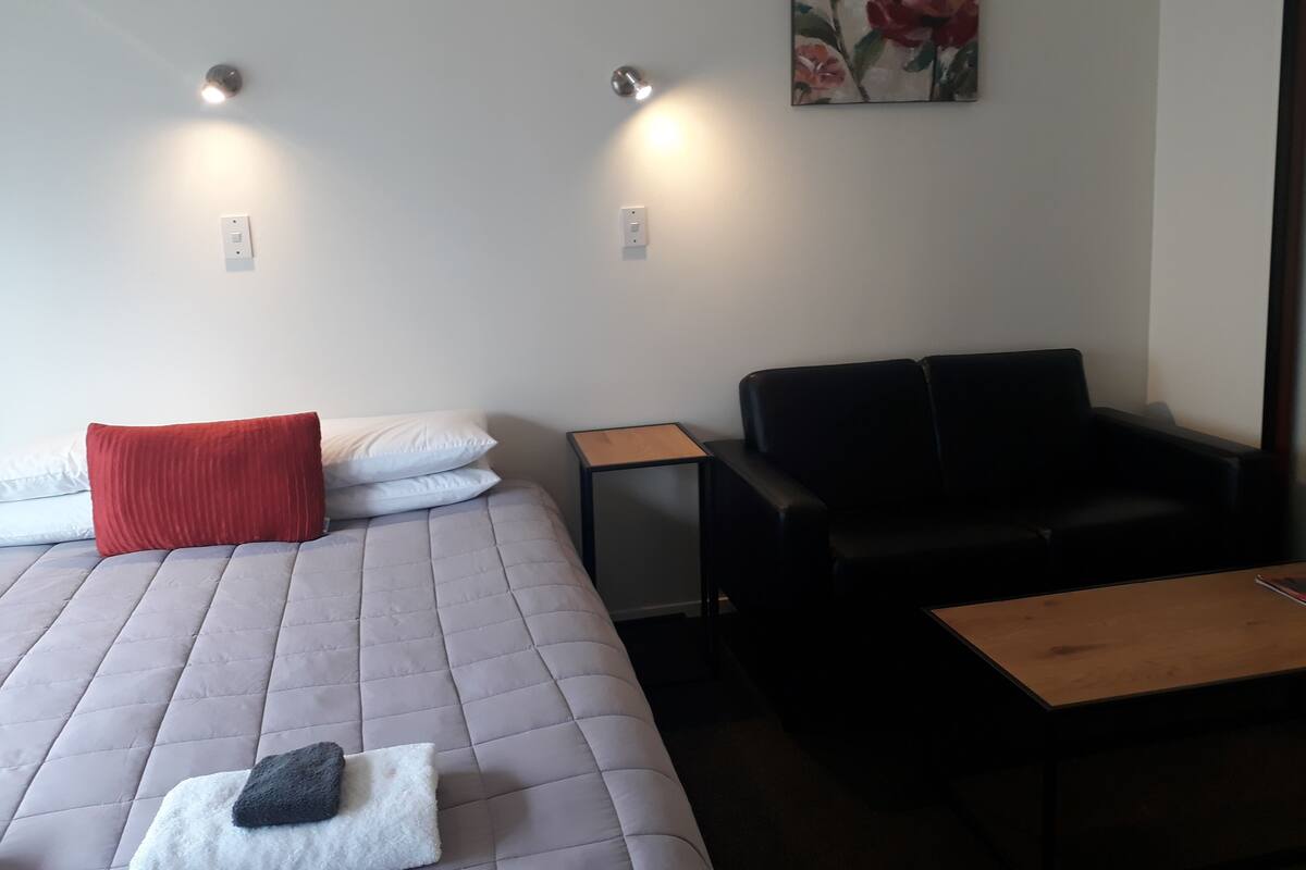 A room with a bed, a black leather sofa, a small wooden table, and a painting on the wall.