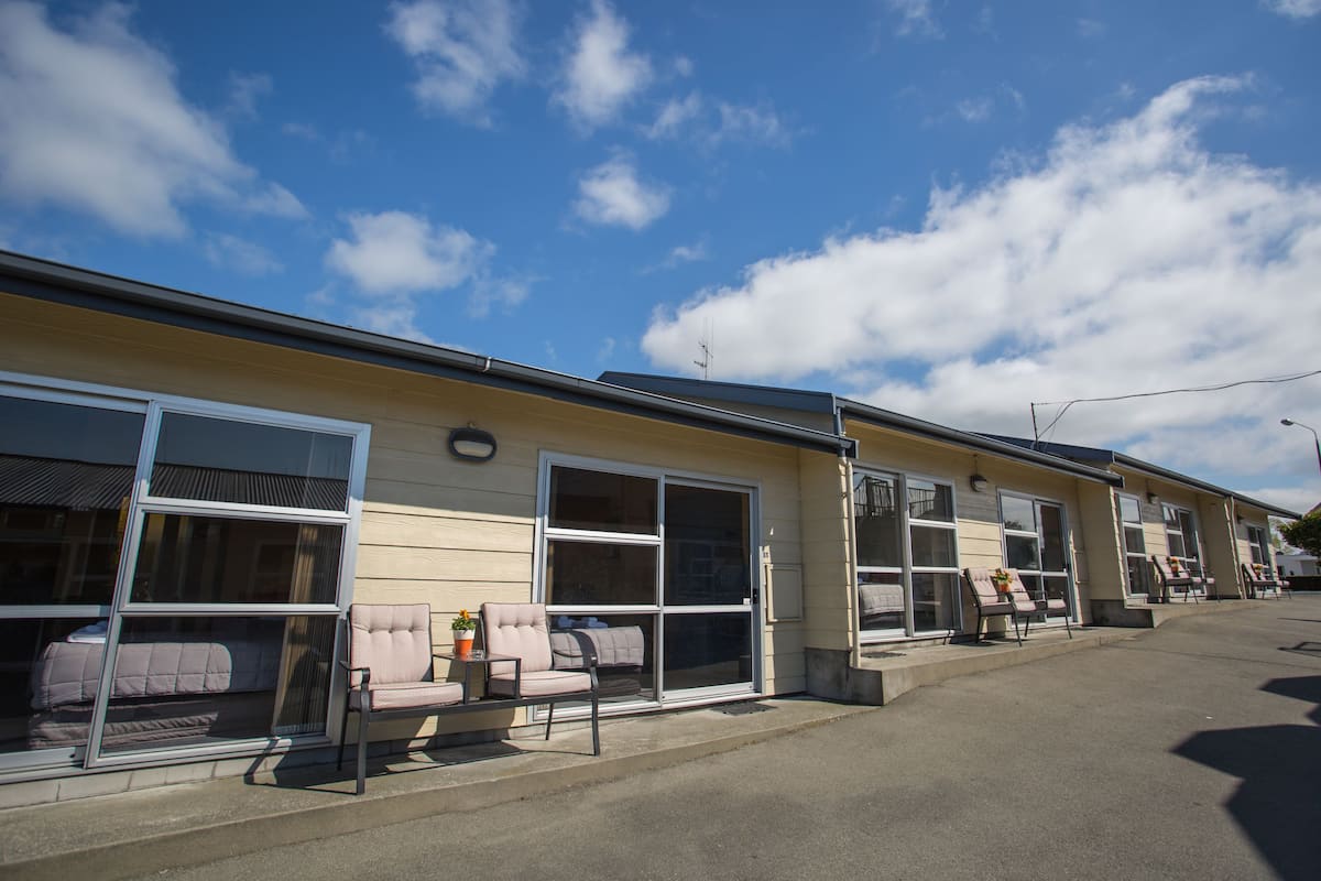 A row of motel rooms with large sliding glass doors, a small outdoor seating area with a chair and table, and a clear blue sky with clouds.