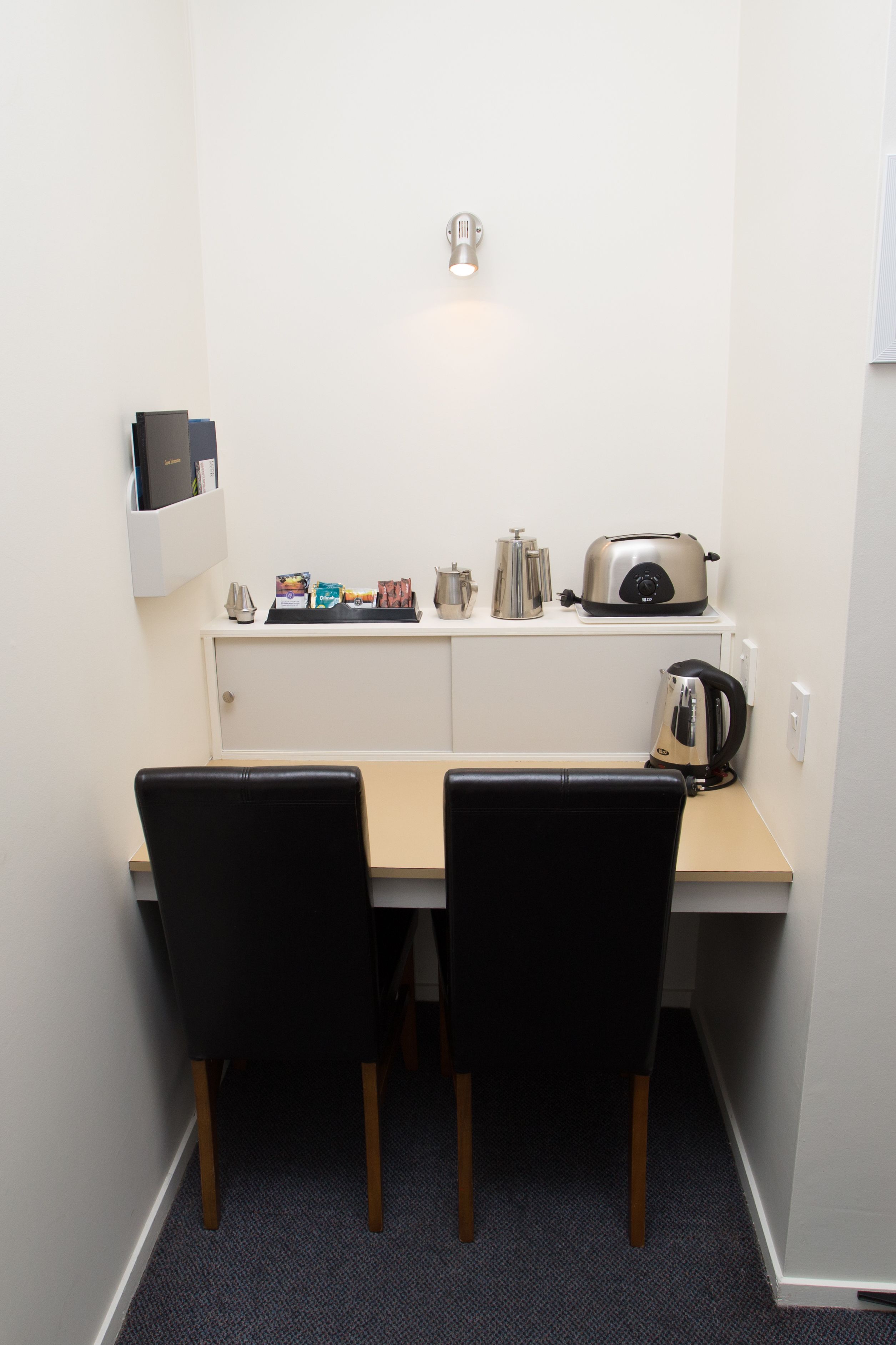 Una pequeña kitchenette con una encimera, un tostador, un hervidor, y una cafetera. Hay dos sillas y un estante con libros y un teléfono.