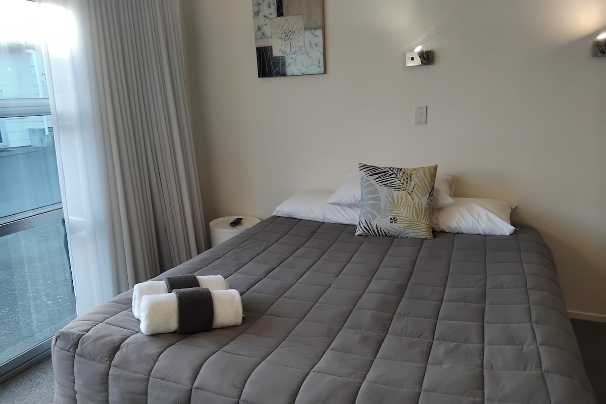 A neatly made bed with a grey quilt, white and patterned pillows, and a pair of folded towels on the footboard.