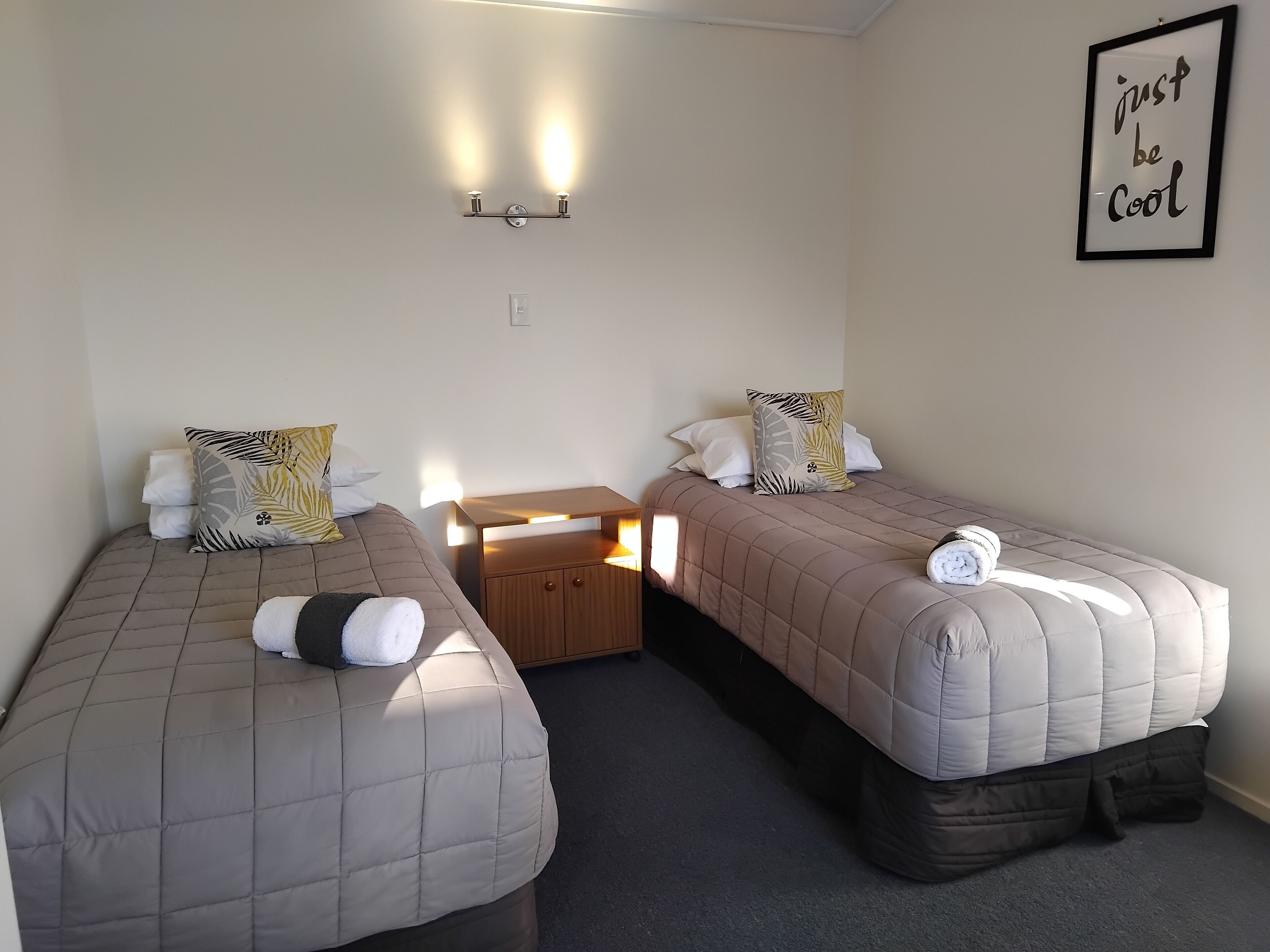 A room with two beds, each with a pillow and a blanket, a small wooden table, and a wall-mounted light fixture.