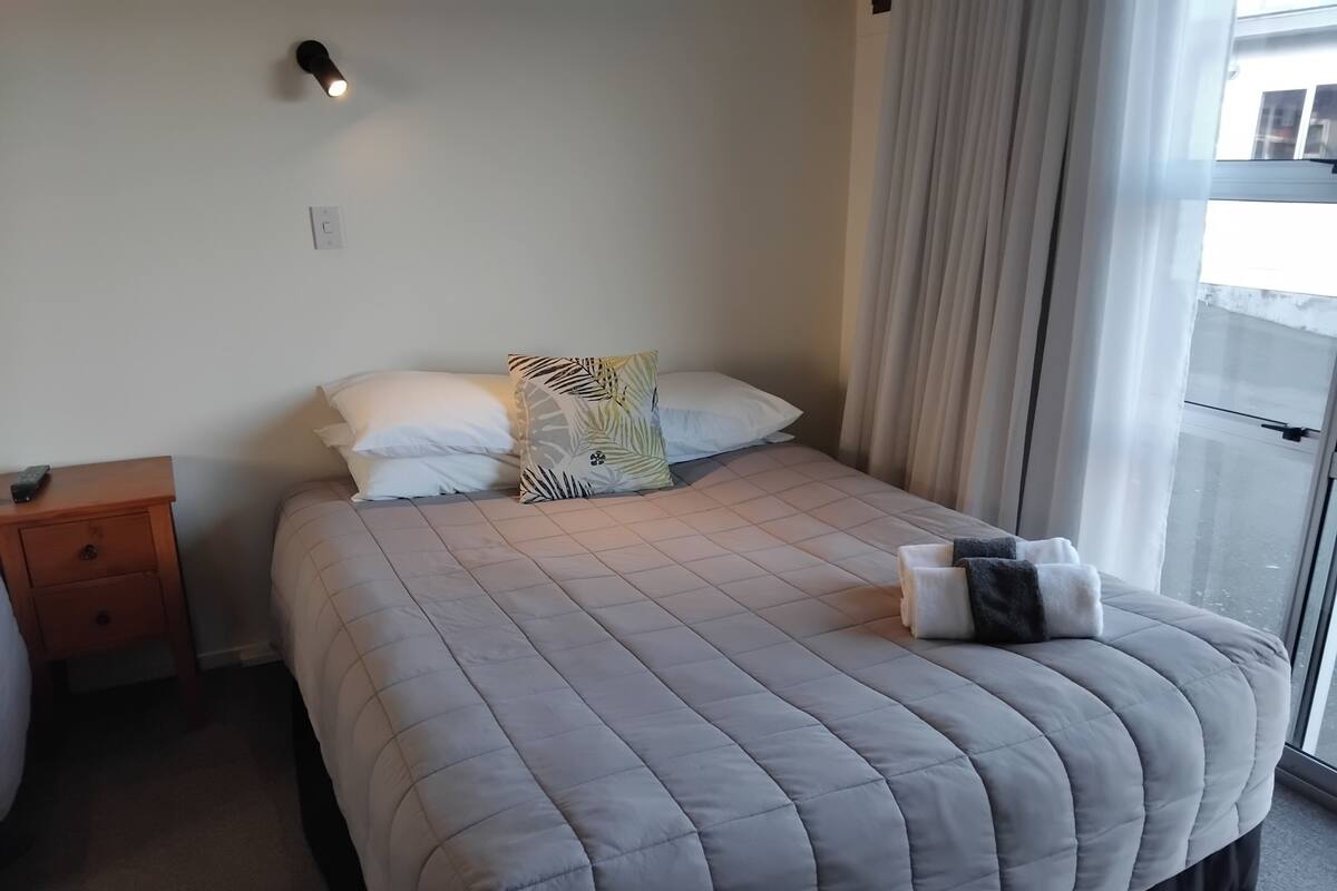 A neatly made bed with a quilted grey duvet, a wooden nightstand with a remote control, and a window with sheer curtains.