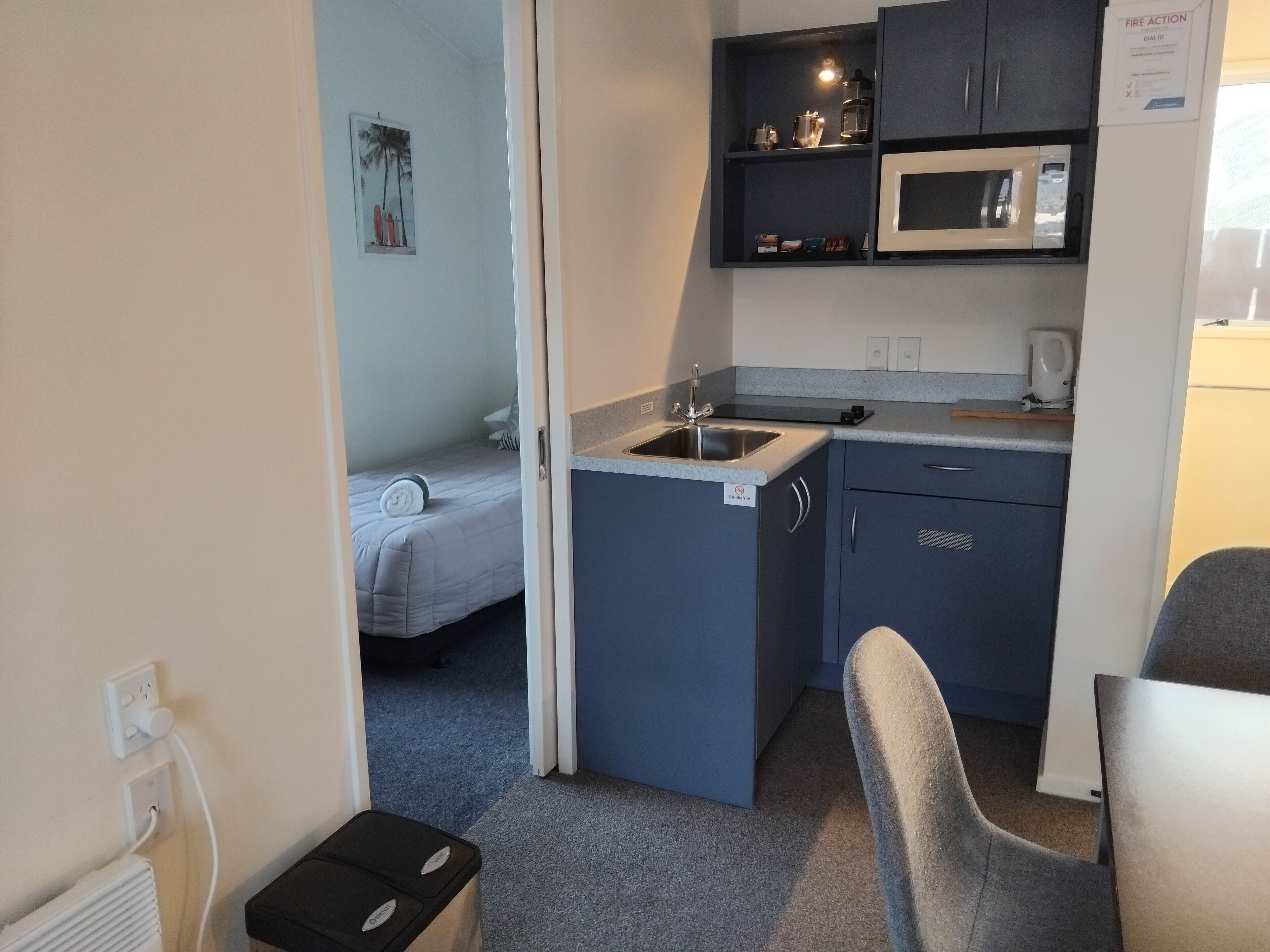 Eine kompakte Küchenzeile mit Mikrowelle, Spüle und Schränken. Ein Bett ist mit einem gerollten Handtuch und ein kleines Regal mit Gegenständen ausgestattet.