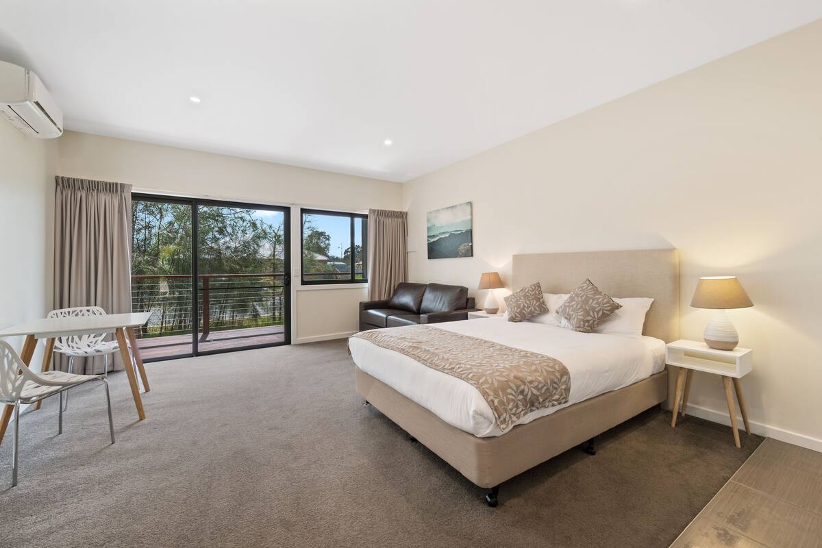 A modern hotel room with a large bed, a desk, and a view of the outdoors through a sliding door.