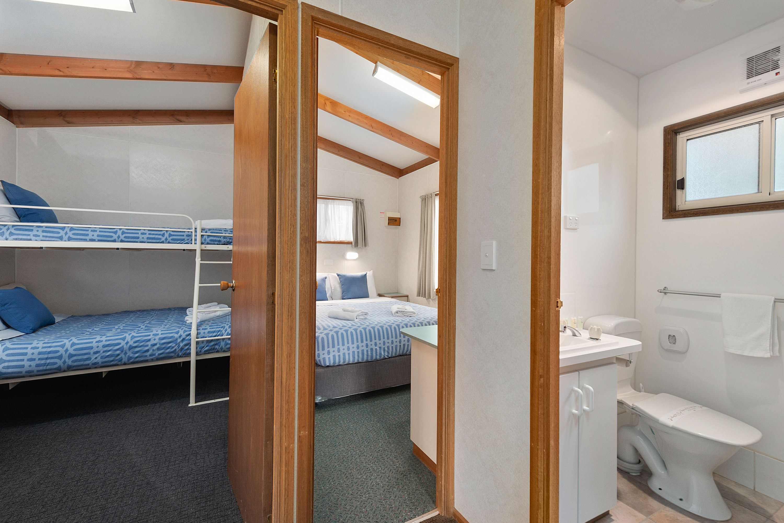 Habitación de hotel pequeña con literas, una cama individual, y un baño compacto con inodoro, lavamanos y espejo.