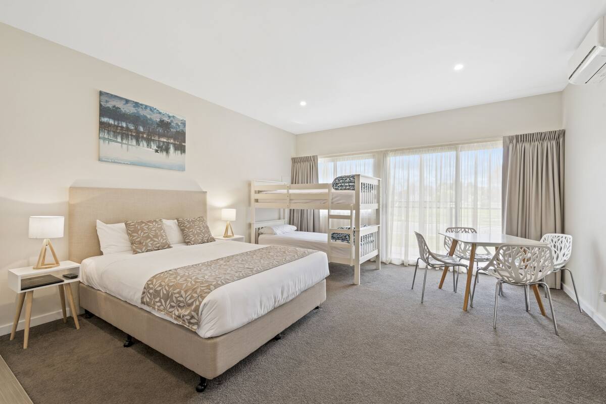 A hotel room with a large bed, a bunk bed, a dining area with chairs, and a painting on the wall.