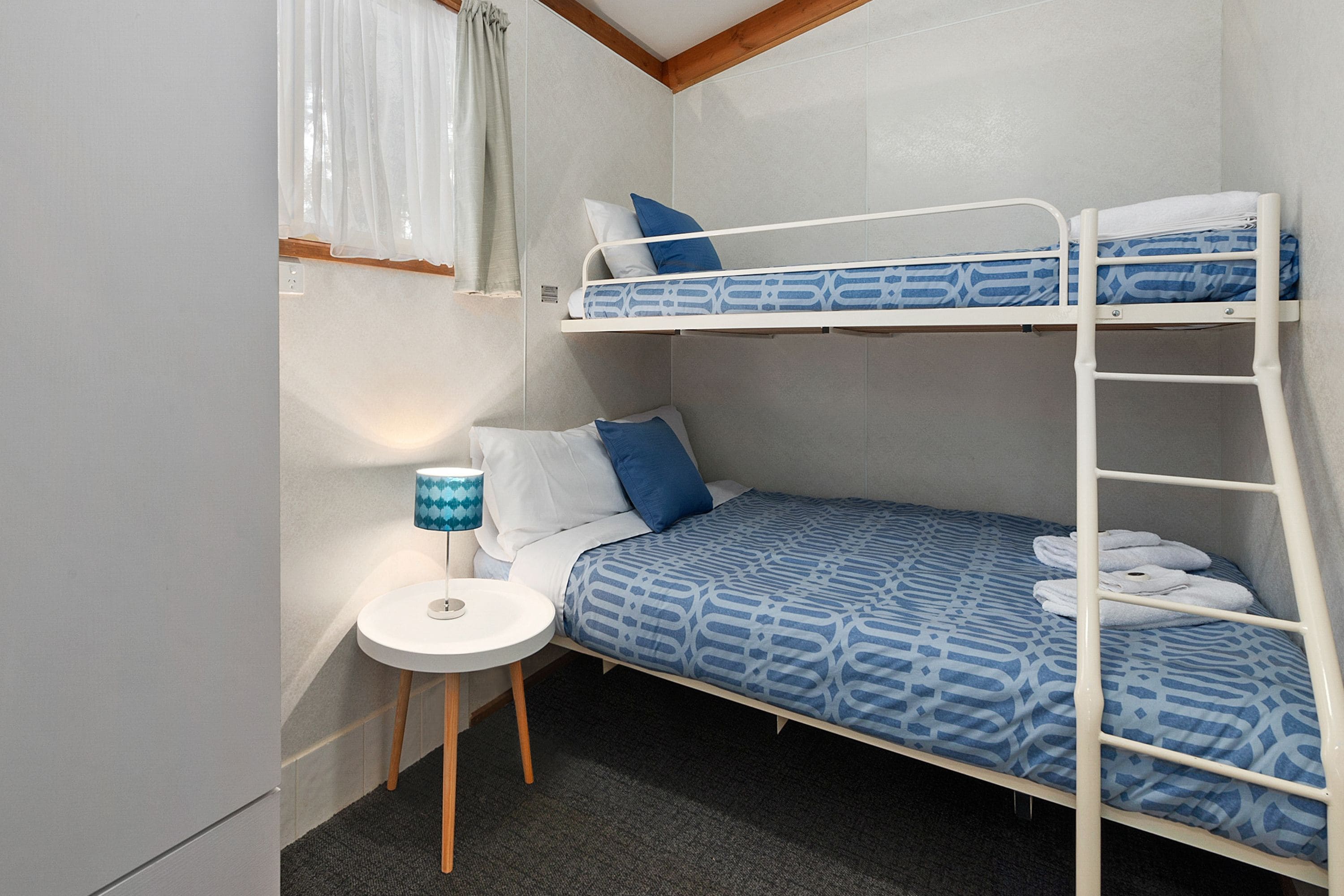 A bunk bed room with a small table and lamp, white walls, and a window with curtains.
