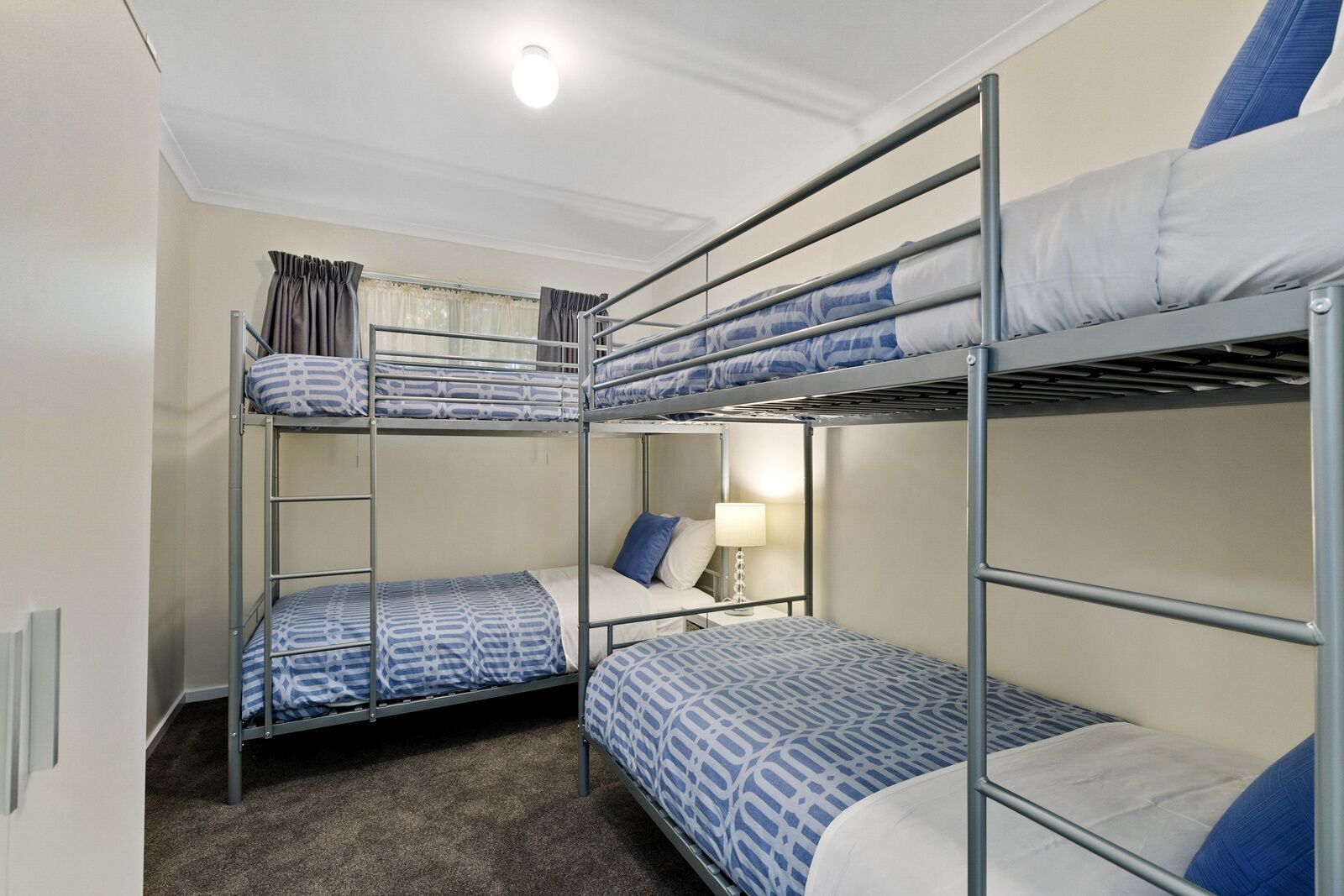 Un dormitorio con literas, tres en total, cada una con ropa de cama estampada, una lámpara de noche y una ventana con cortinas.