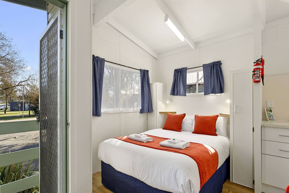 A hotel room with a large bed, a bedside table, a nightstand, a window with curtains, a fire extinguisher, and a view of the outside.