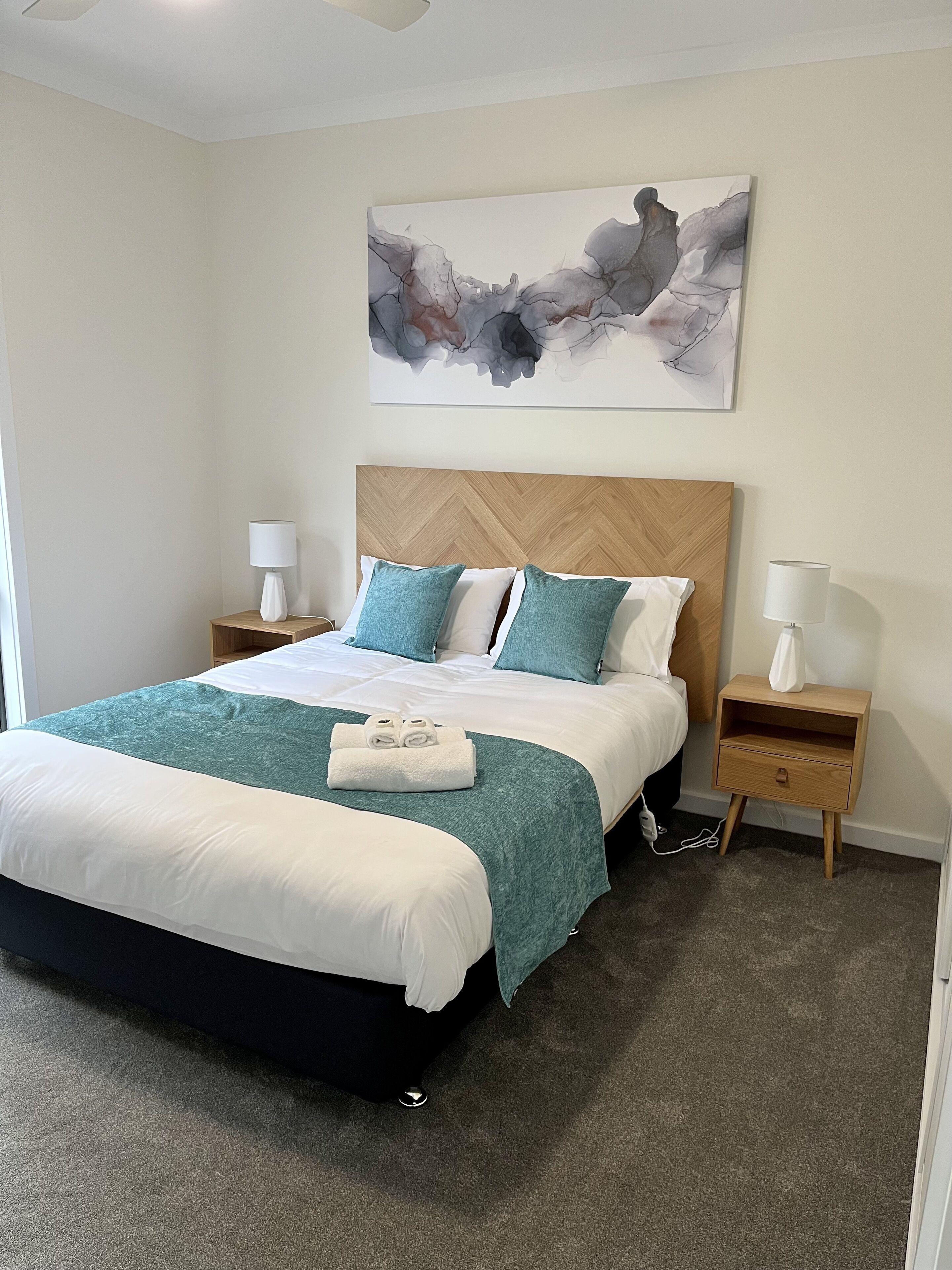 A neatly arranged bedroom with a large bed, two bedside tables, a wall-mounted abstract painting, and a carpeted floor.