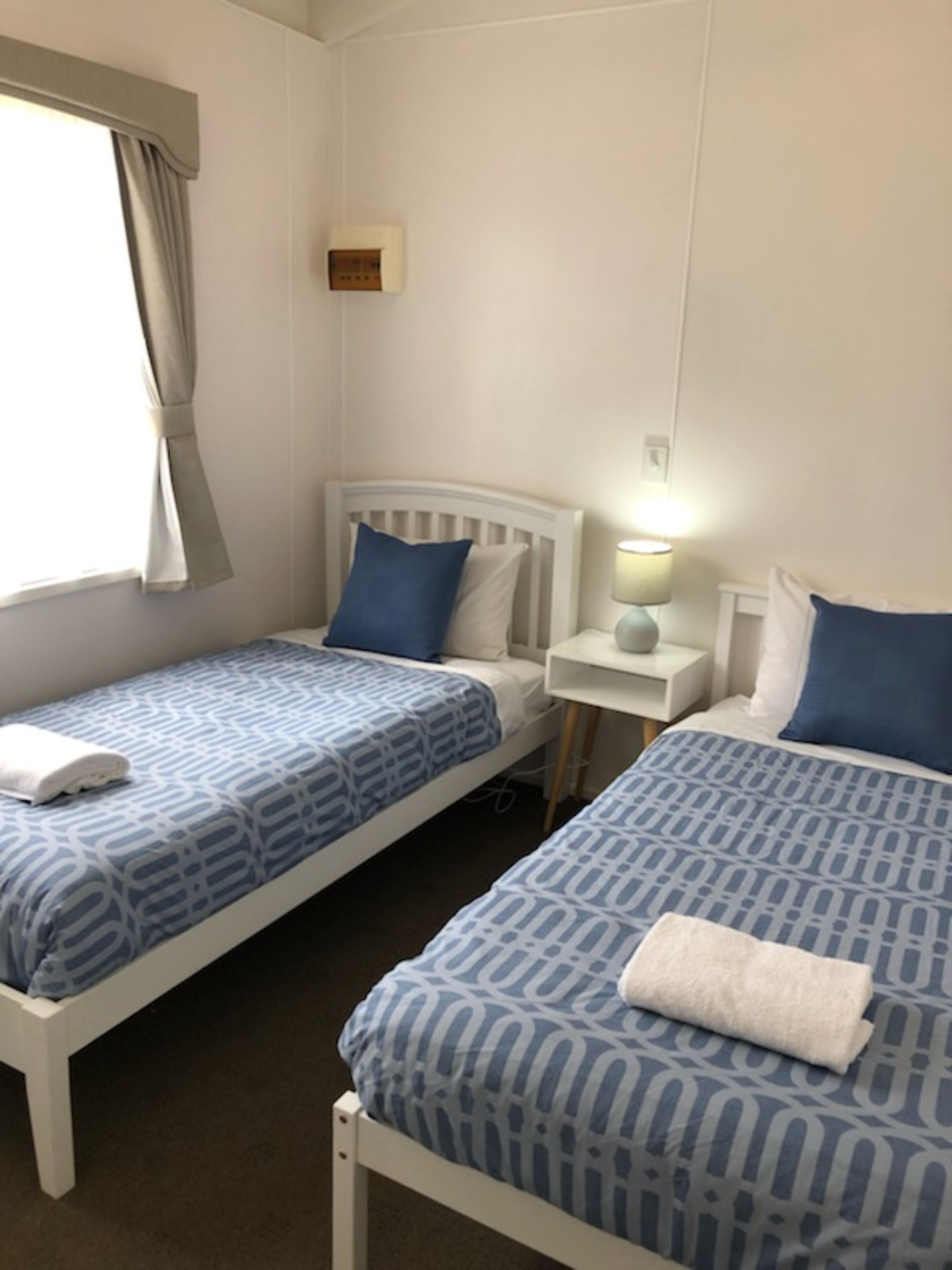 A hotel room with two single beds, each with a blue patterned bedspread and pillows, a bedside table with a lamp, and a window with curtains.