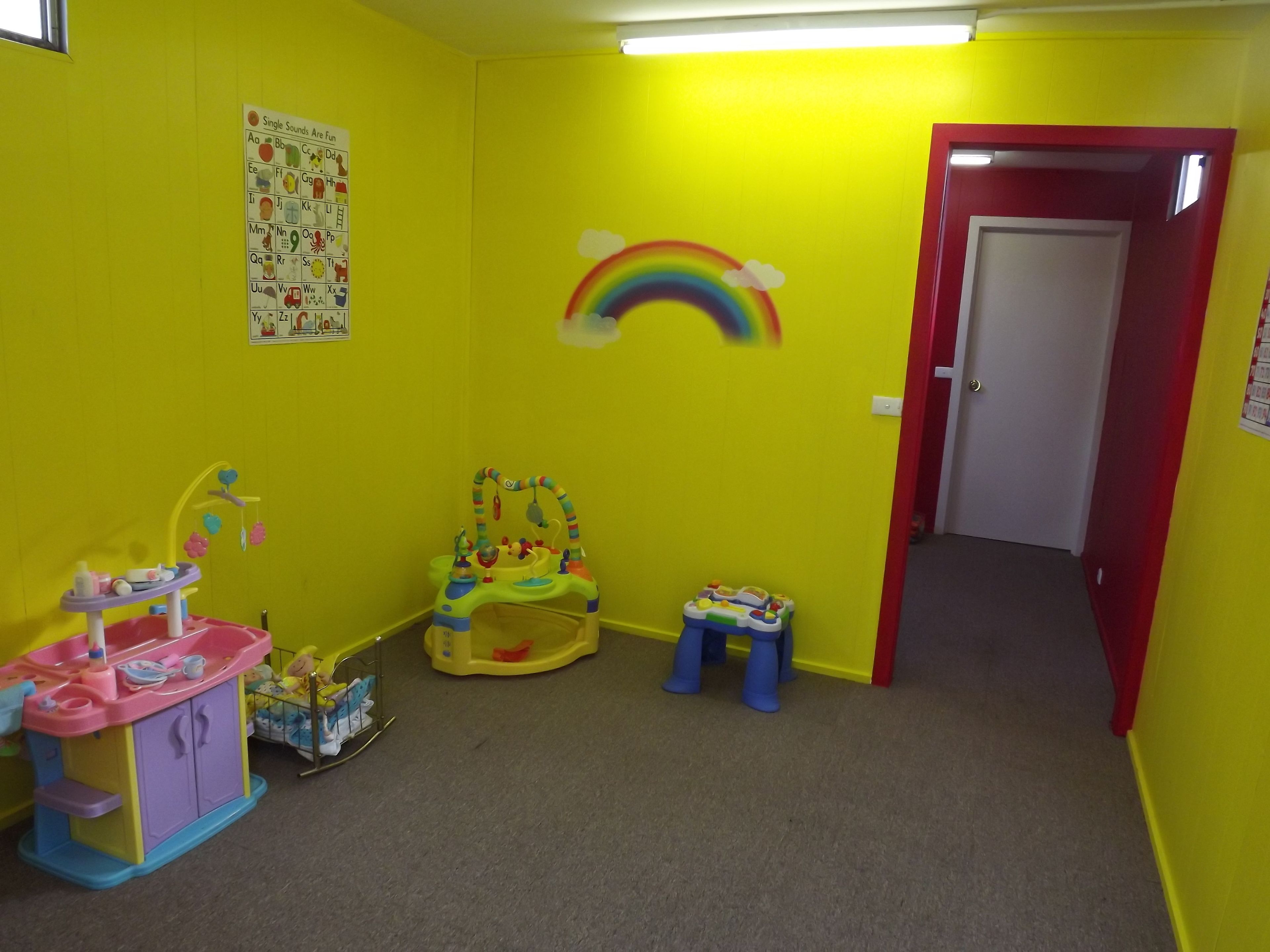 Un salón infantil lleno de color, con un mural de arcoíris, un corralito y una cocina de juguete.