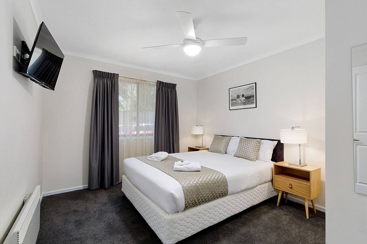 A hotel room with a bed, a TV mounted on the wall, a ceiling fan, a bedside table with a lamp, and a framed picture on the wall.