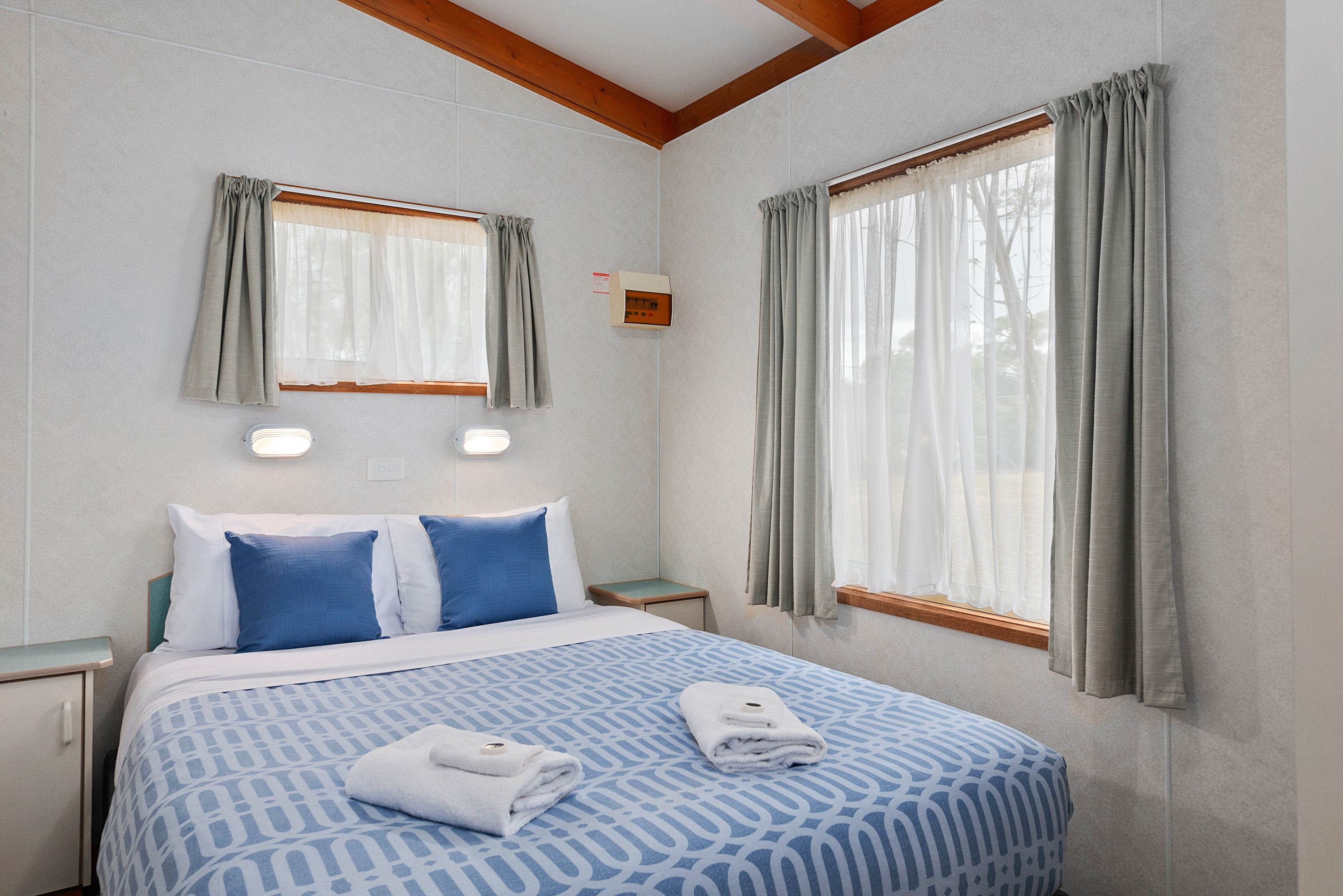 A hotel room with a bed, two bedside tables, a window with curtains, and a thermostat on the wall.