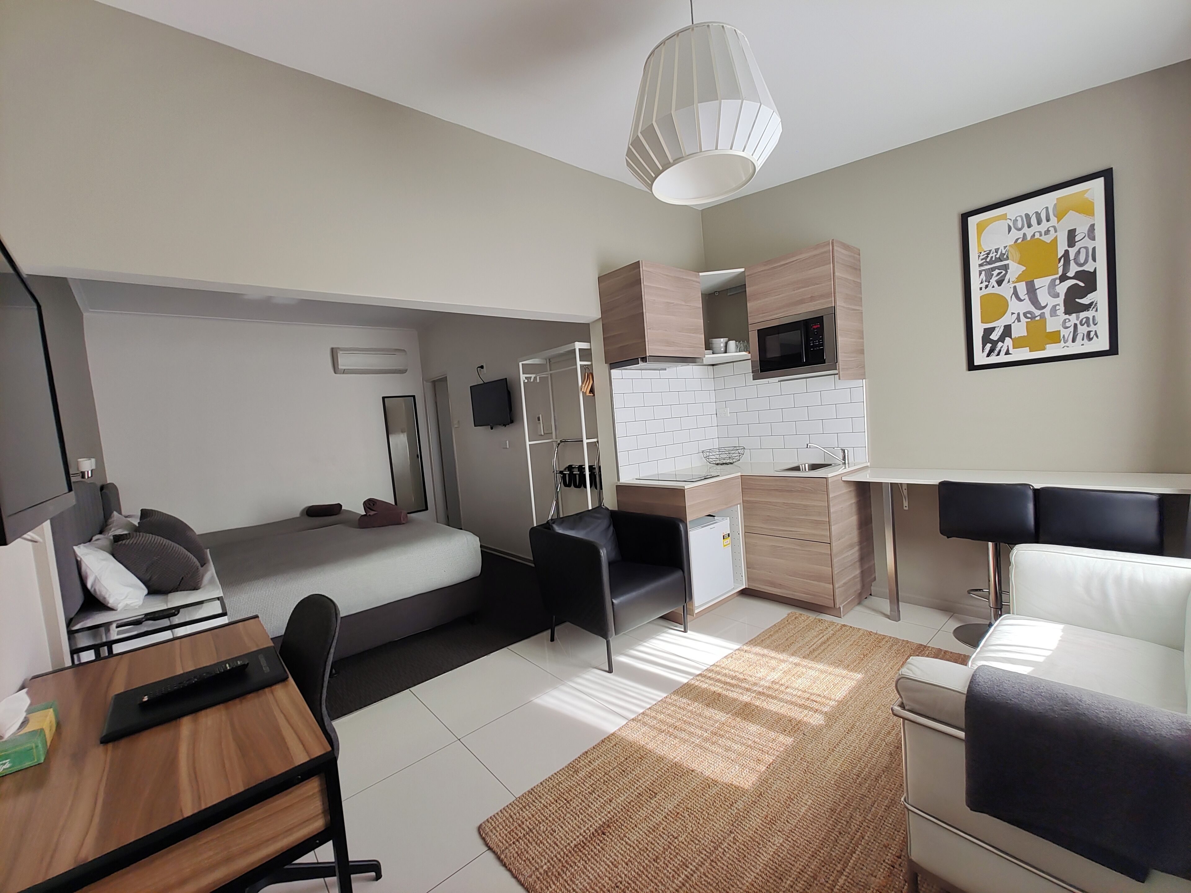 A compact hotel room with a bed, a desk, a kitchenette, and a dining area.