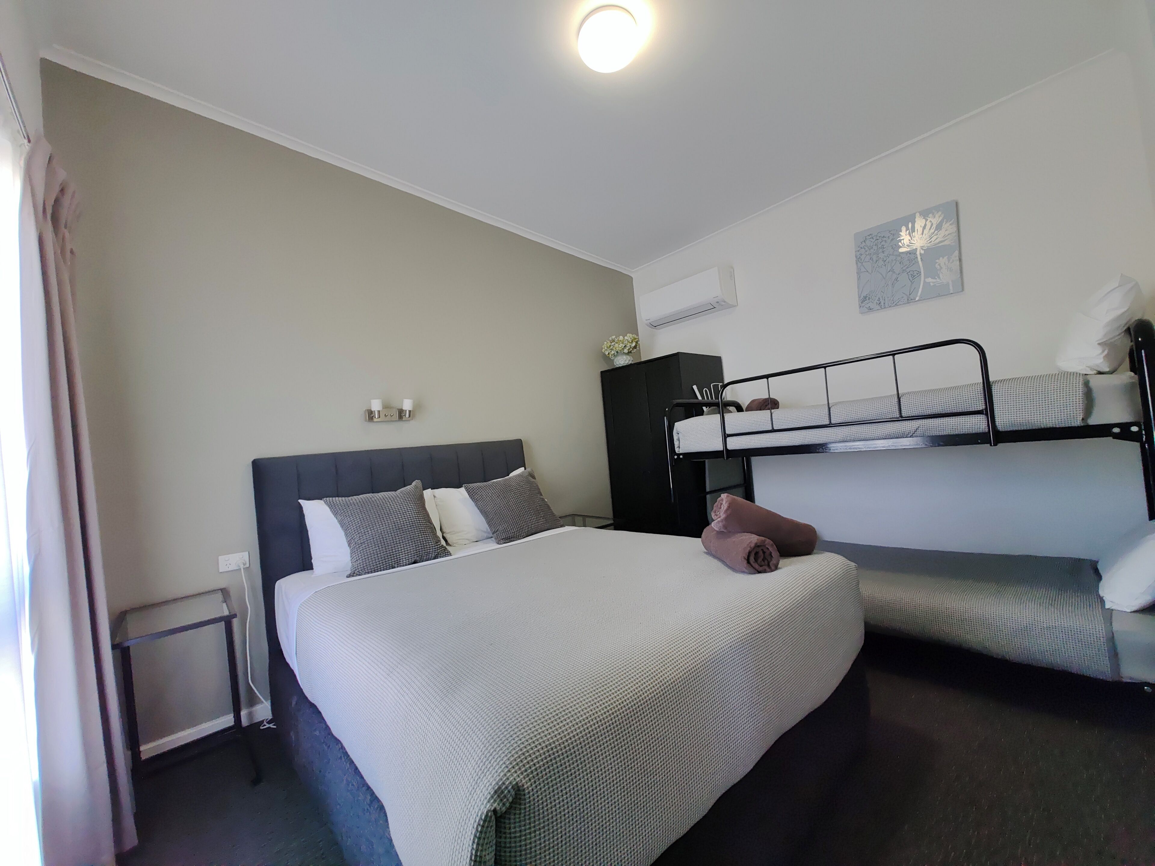 A hotel room with a single bed, a bunk bed, a small table, and a wall-mounted air conditioner.