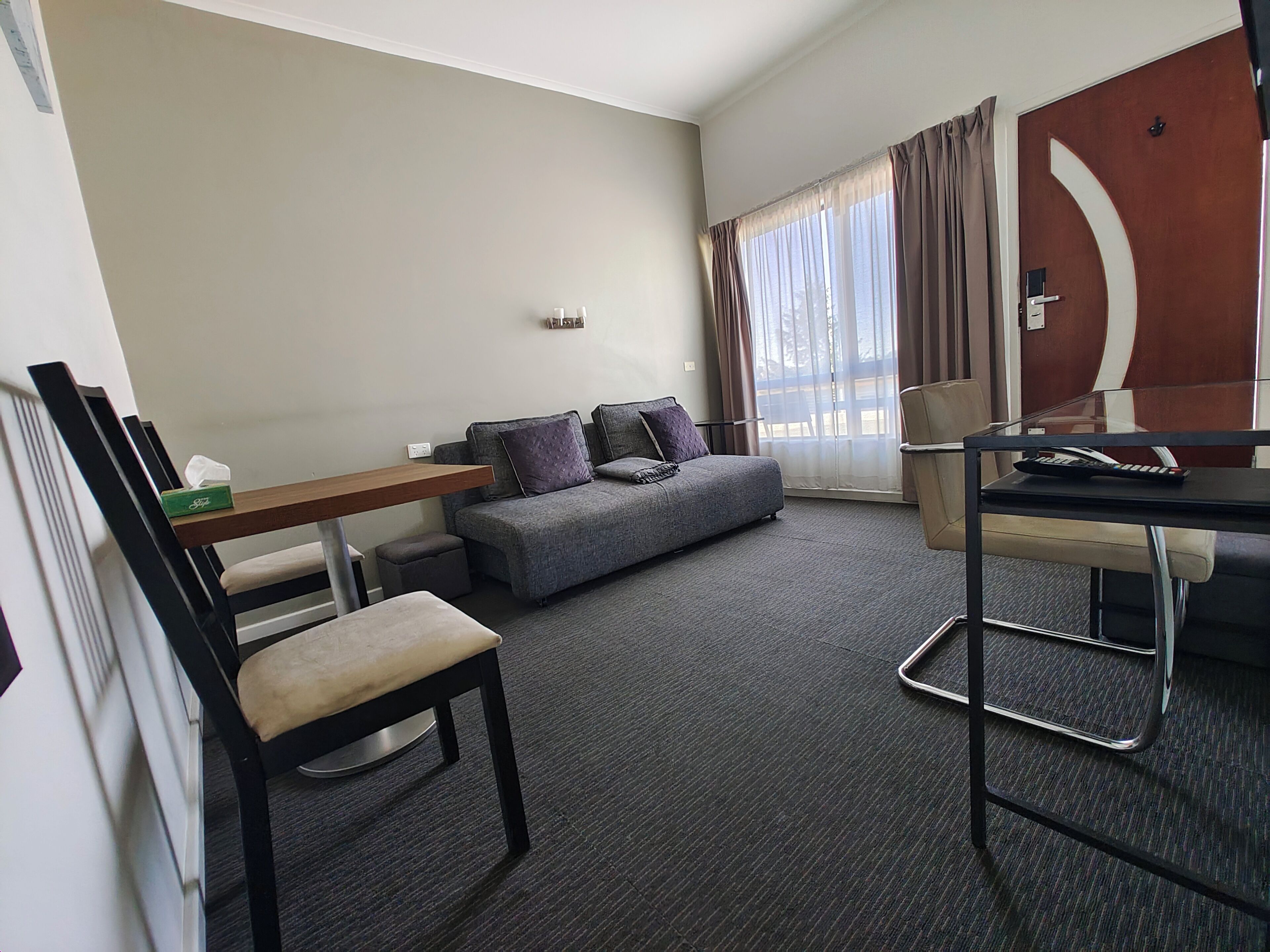 A modern hotel room with a grey sofa, a wooden desk, a chair, and a glass table.