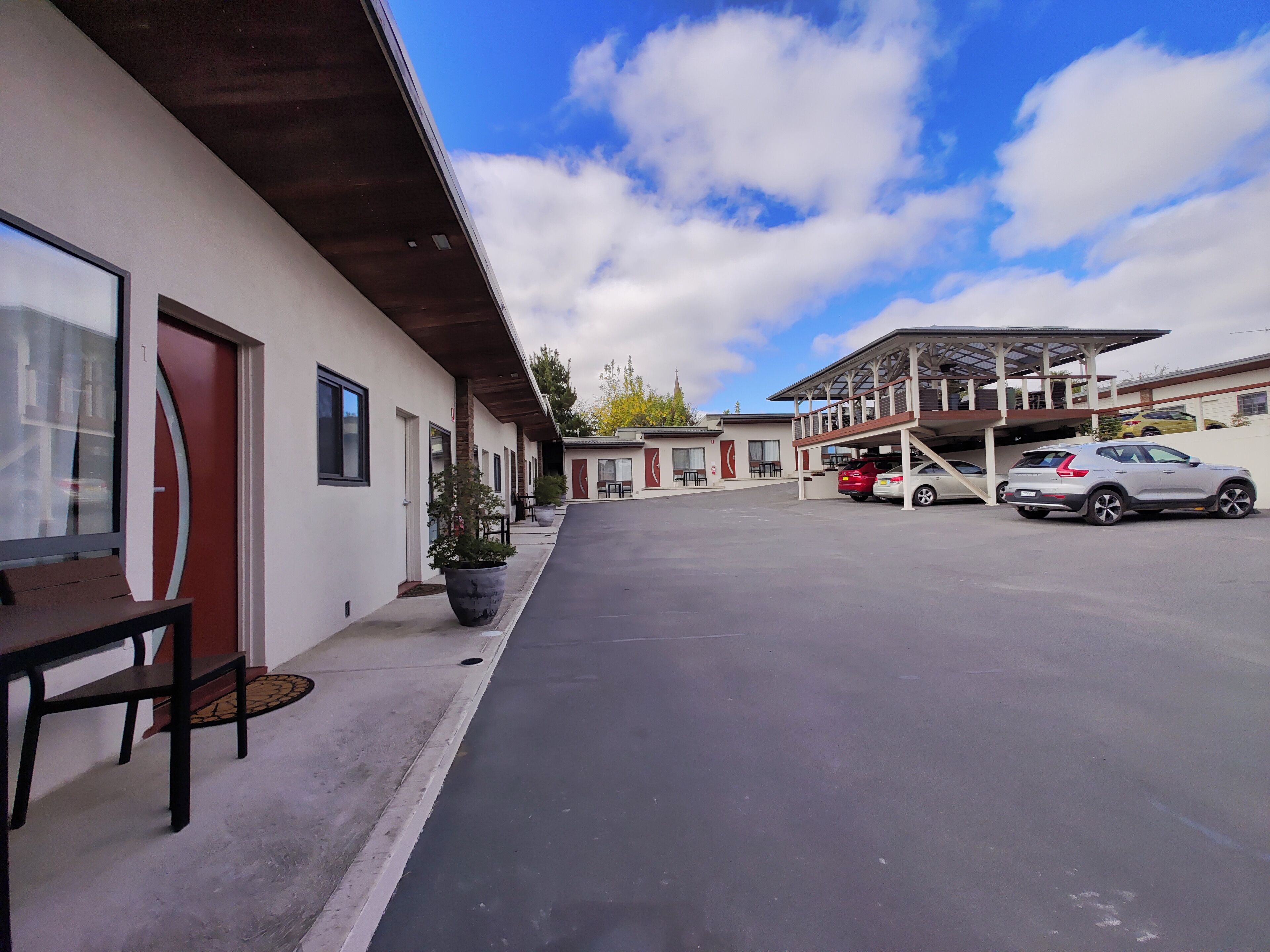 A hotel with a covered outdoor seating area, a parking lot, and a building with a sloped roof.