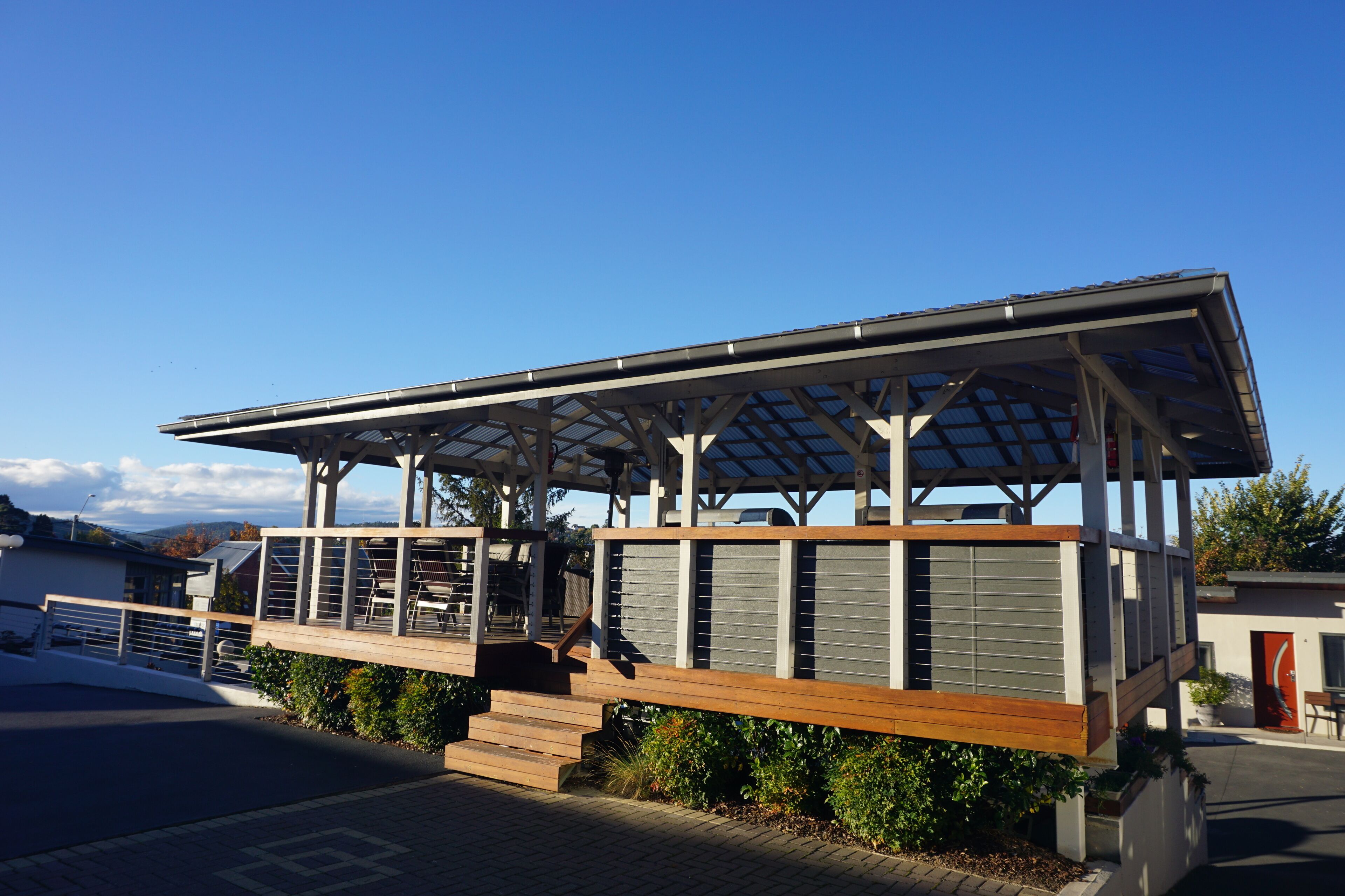 A covered outdoor seating area with a wooden deck and metal railings, adjacent to a building with a sloped roof and large windows.
