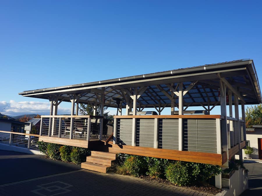 Un espace extérieur abrité, doté d’une terrasse en bois et de rampes en métal, jouxtant un bâtiment avec un toit en pente et de grandes fenêtres.