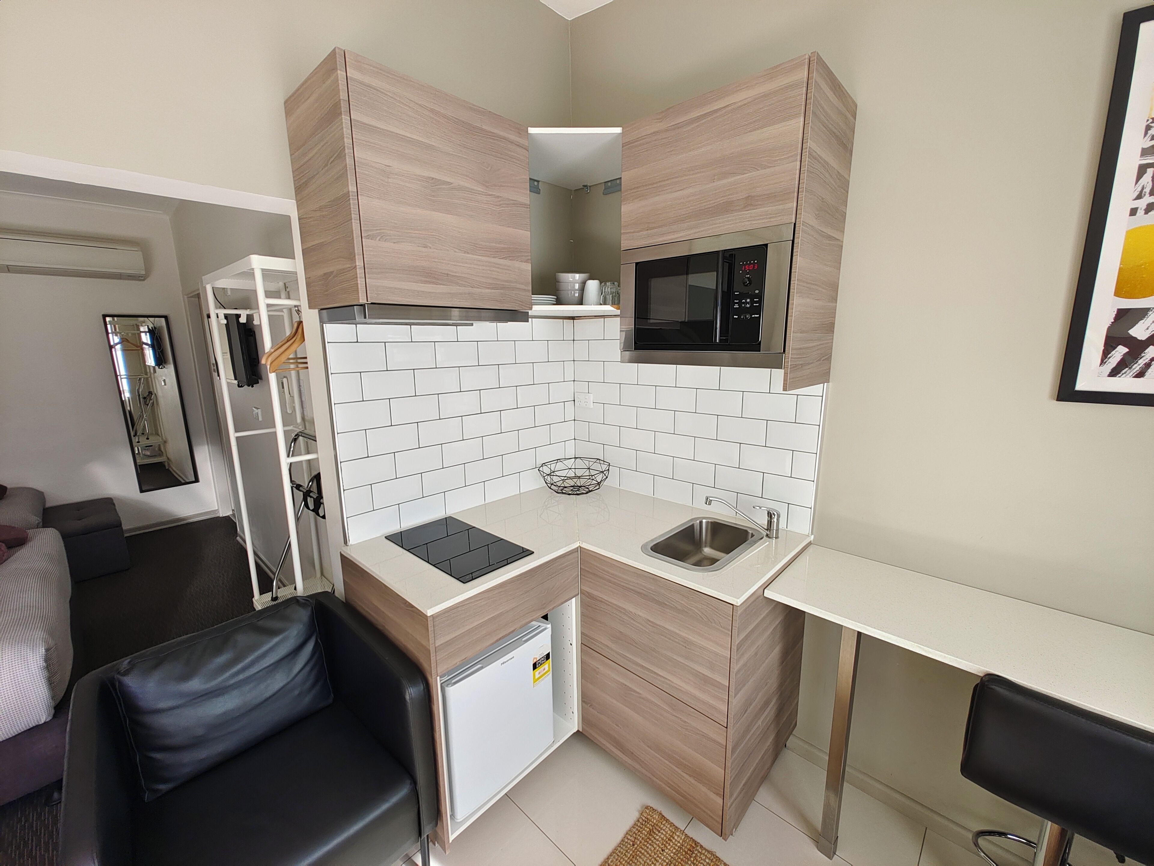 Una cocina compacta con microondas, lavamanos y un refrigerador pequeño. Hay un armario de madera sobre la encimera y un espejo en la pared.