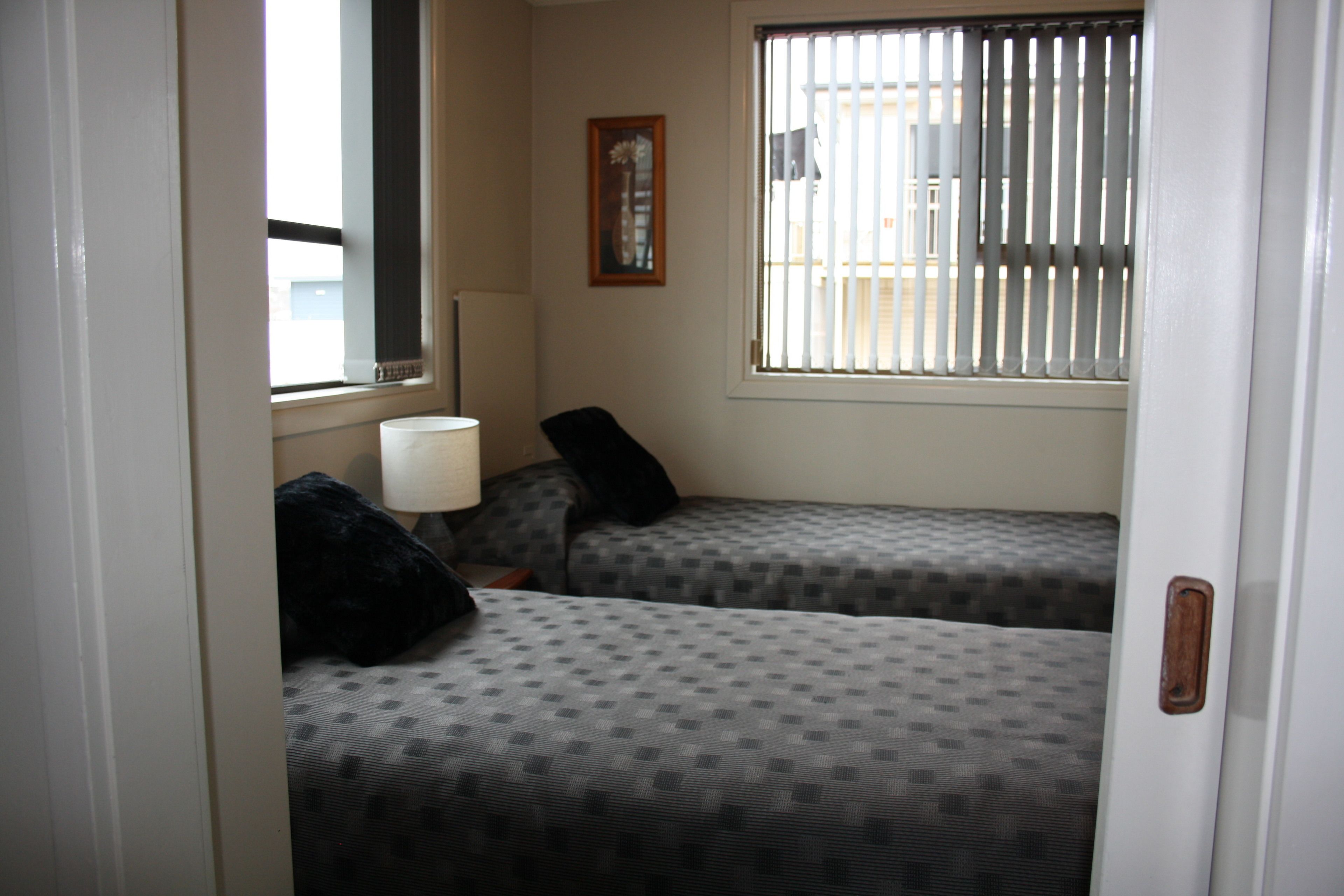 A small room with two beds, a window with blinds, a lamp, and a framed picture on the wall.