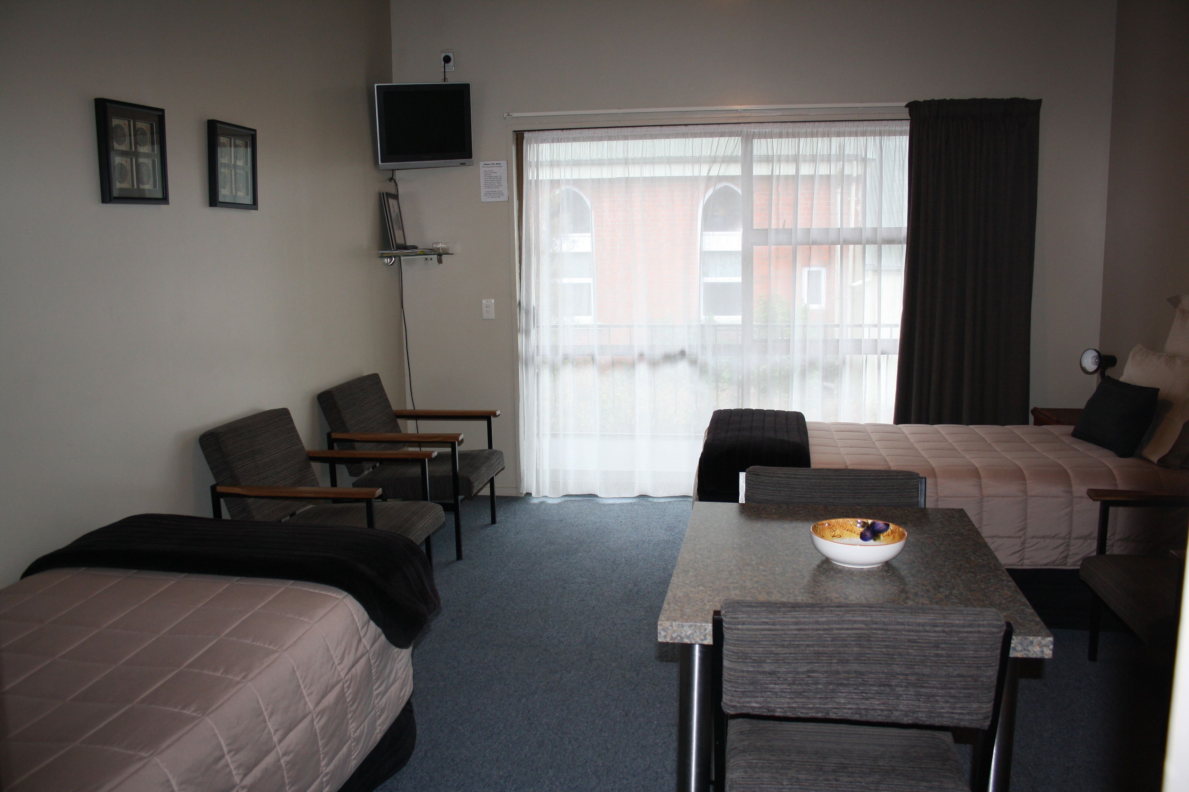 Habitación de hotel con una cama, dos sillas, una mesa con un cuenco, un televisor en la pared y una ventana con cortinas.