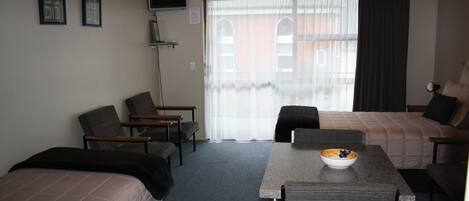 Habitación de hotel con una cama, dos sillas, una mesa con un cuenco, un televisor en la pared y una ventana con cortinas.