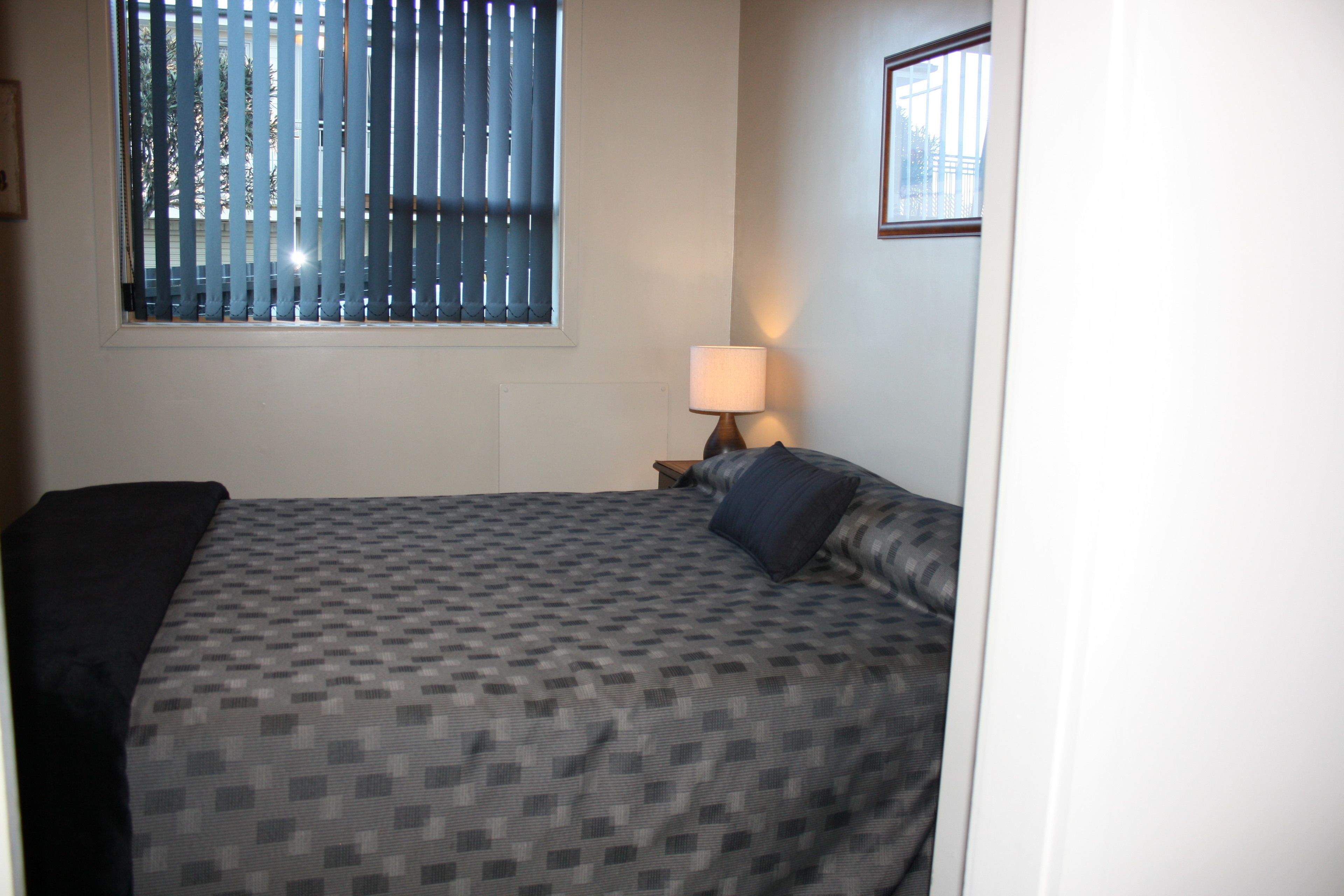 Une chambre d’hôtel avec un lit, une fenêtre avec des stores, une table de chevet avec une lampe et un tableau encadré au mur.