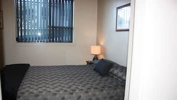 A hotel room with a bed, a window with blinds, a nightstand with a lamp, and a framed picture on the wall.