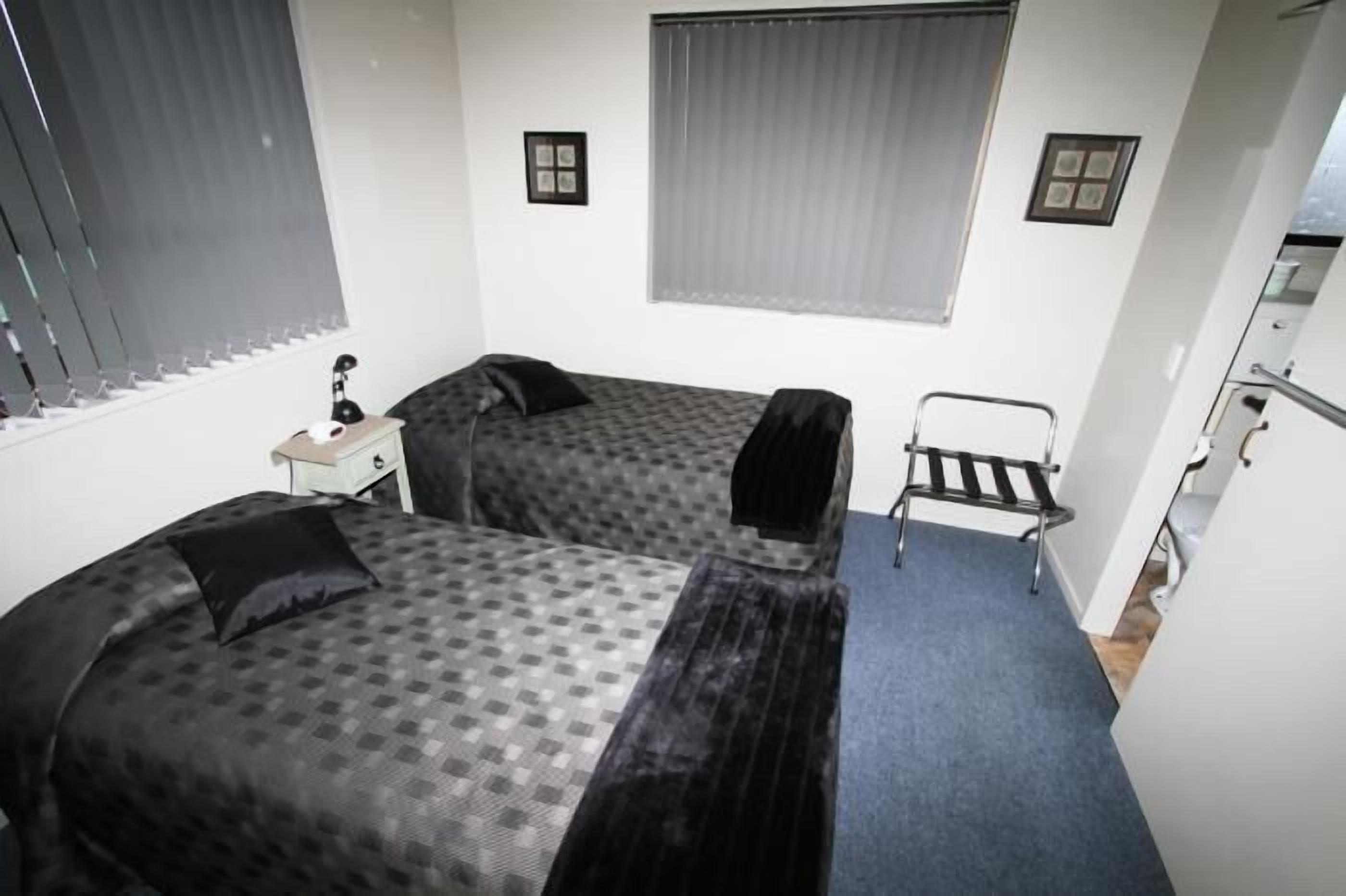 A small, compact room with two beds, a desk, a chair, and a window with blinds.