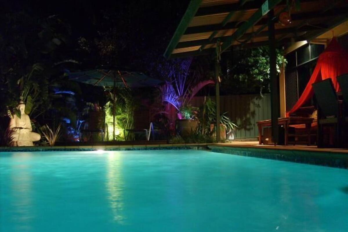 A nighttime pool area with a statue, potted plants, and a covered seating area.