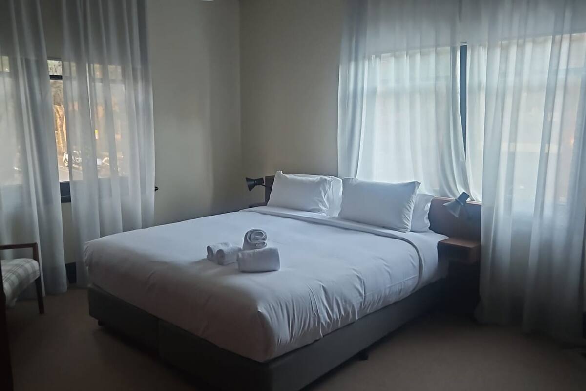 A neatly made bed with white linens and a folded towel, a bedside table with a lamp, and sheer curtains on the windows.