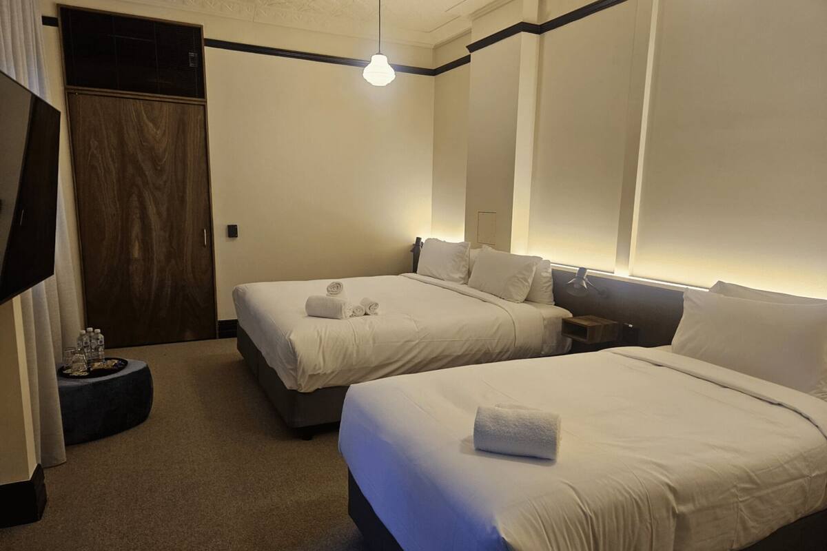 A hotel room with two beds, a wooden door, a small round table with water bottles, and a hanging light fixture.
