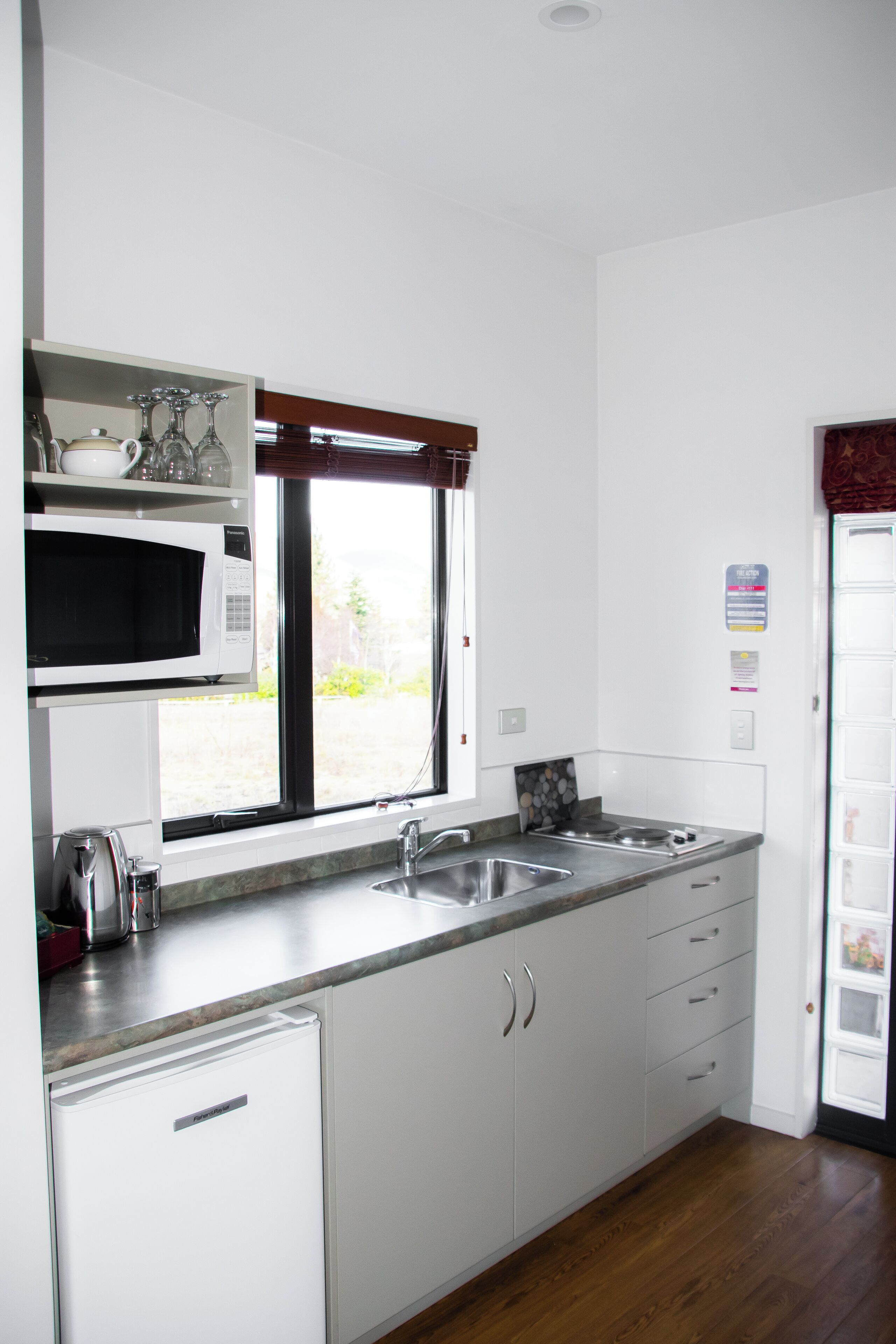 Una cocina moderna con armarios blancos, un microondas empotrado, un lavamanos de acero inoxidable y una ventana con persianas.
