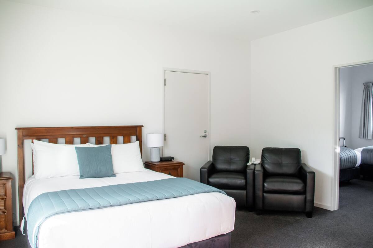 A hotel room with a bed, two chairs, a nightstand, and a door.