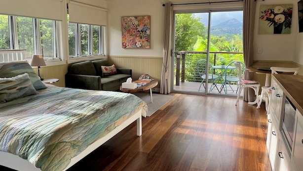 Un dormitorio con cama, sofá, zona de comedor y vistas a la vegetación a través de amplias ventanas.
