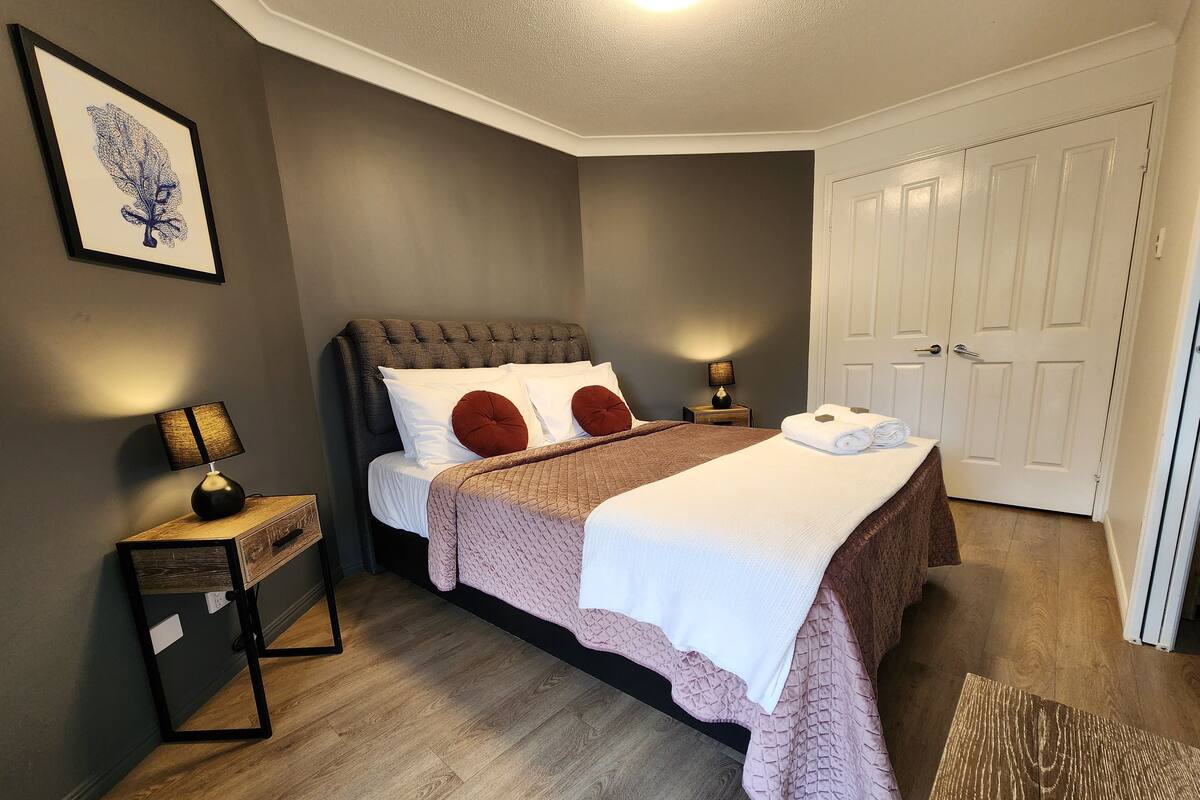 A neatly arranged bedroom with a bed, bedside lamps, a nightstand, a door, and a framed artwork on the wall.