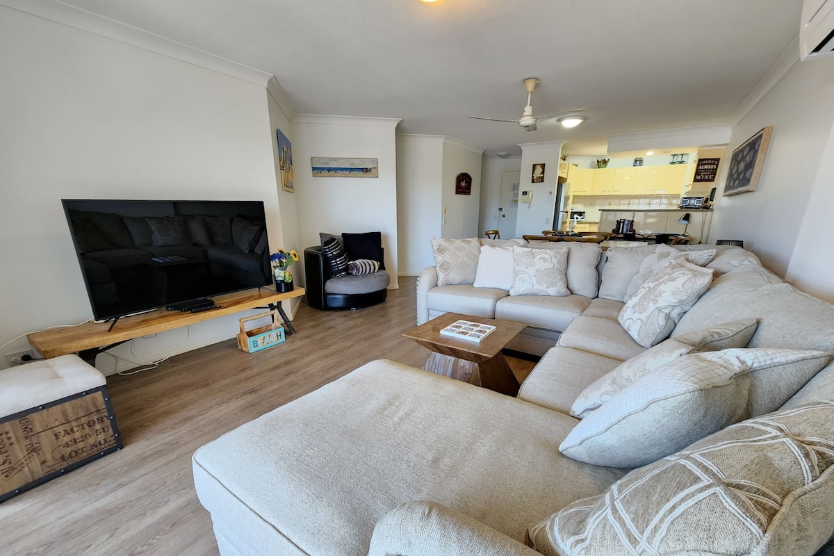 A modern living room with a large flat-screen TV, a beige sectional sofa, a wooden coffee table, and a small side table.