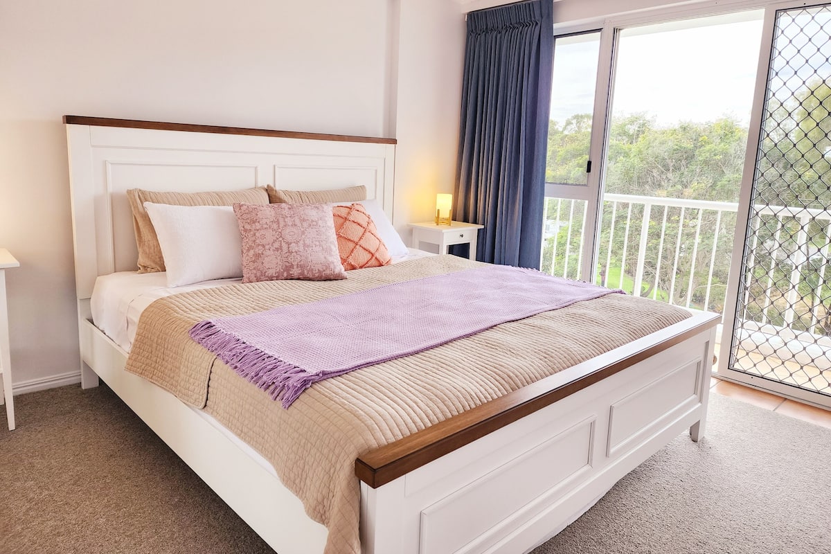 A bedroom with a bed, nightstand, and a view of the outdoors.