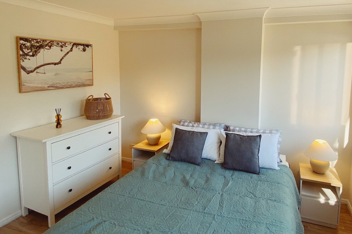 A bedroom with a bed, a dresser, two bedside tables with lamps, a painting on the wall, and a window with curtains.