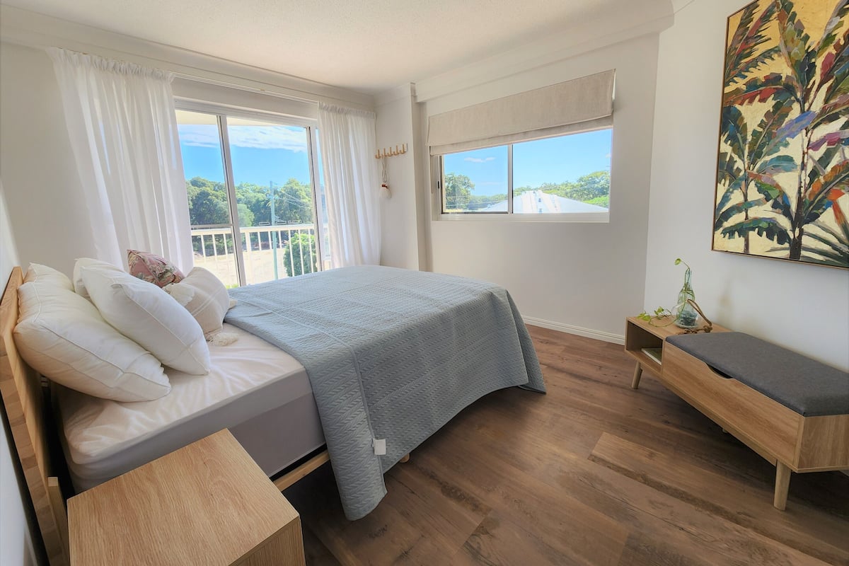 A bedroom with a bed, a bench, a window with a view, and a painting on the wall.