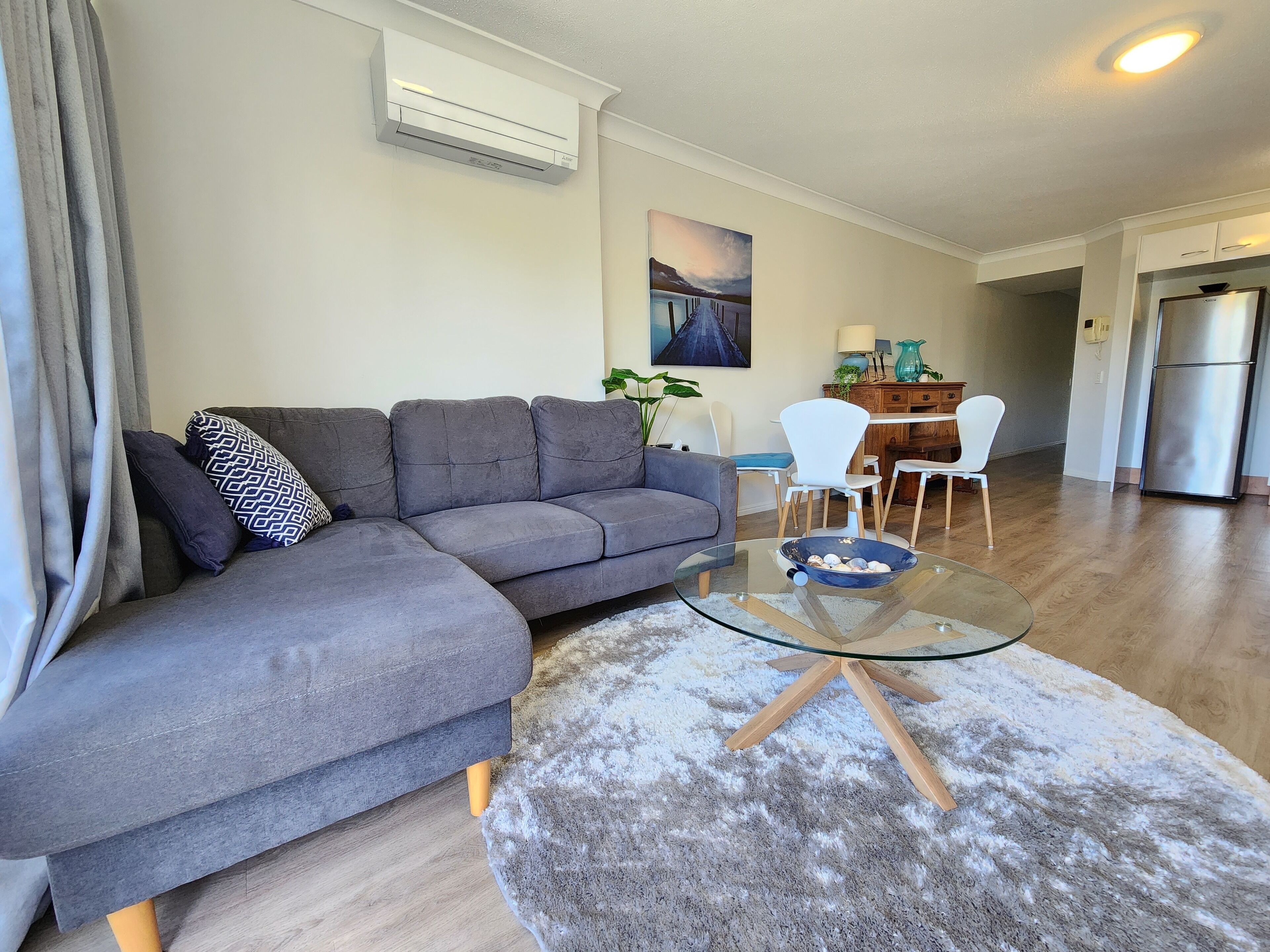 A modern living room with a grey sectional sofa, a glass coffee table, and a dining area with white chairs.