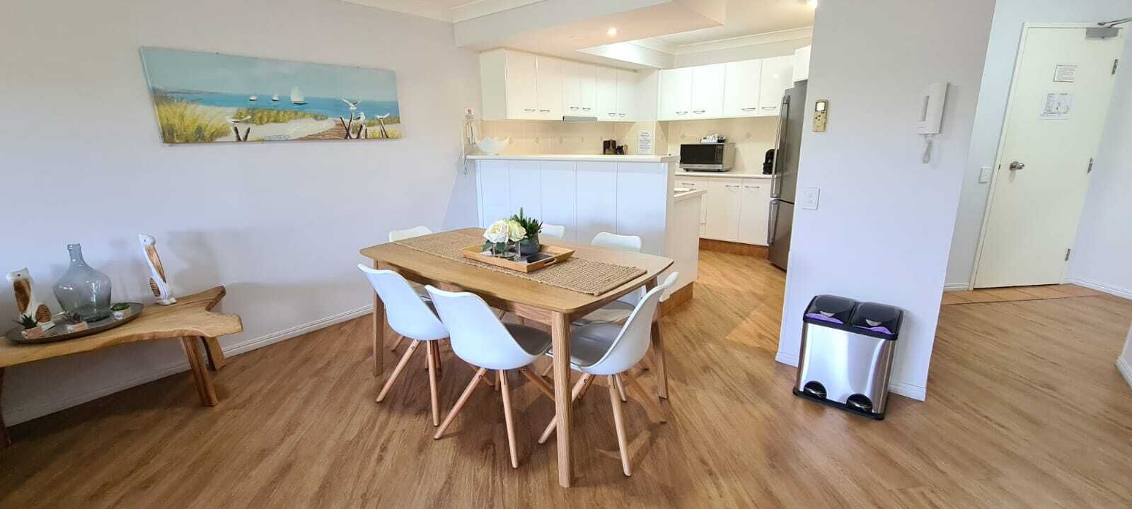 Une cuisine moderne avec des meubles blancs, une table à manger en bois et quatre chaises, ainsi qu’une poubelle.