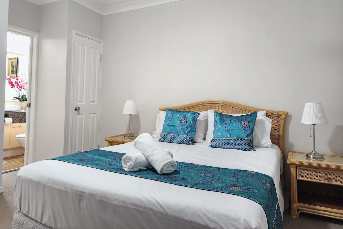 A neatly made bed with white linens and blue patterned bedspread, flanked by two bedside tables with lamps, in a room with white walls and a door leading to a bathroom.