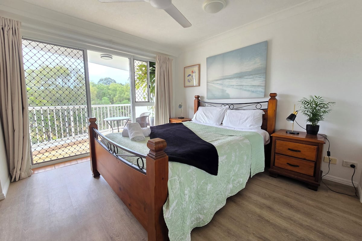 A bedroom with a bed, a nightstand, a potted plant, a painting, and a balcony with a view of greenery.
