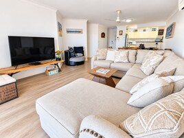 Una sala de estar moderna con un televisor de pantalla plana grande, un conjunto de sofás, una mesa de centro y una zona de comedor con una cocina al fondo.