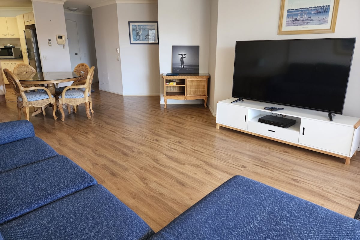 A living room with a flat-screen TV, blue sofas, a dining area with wicker chairs, and a kitchen in the background.