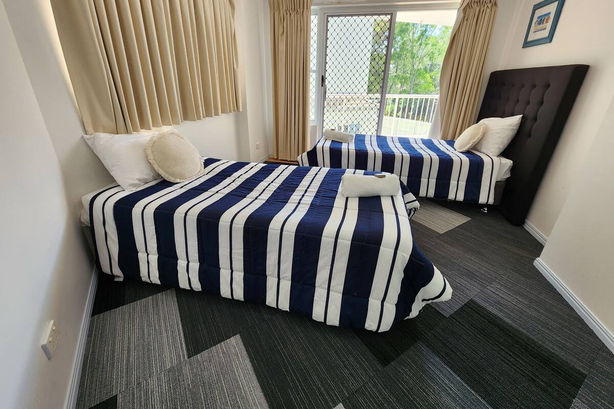 A hotel room with two beds, each with striped bedding, a dark carpet, and a view of the outdoors.