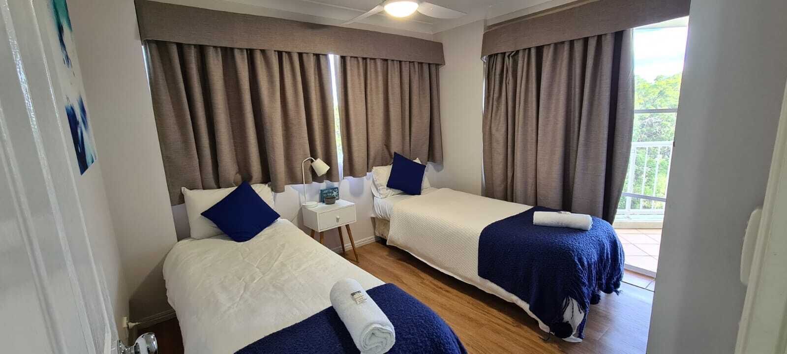 A hotel room with two single beds, a nightstand, a lamp, and a view of the outdoors.